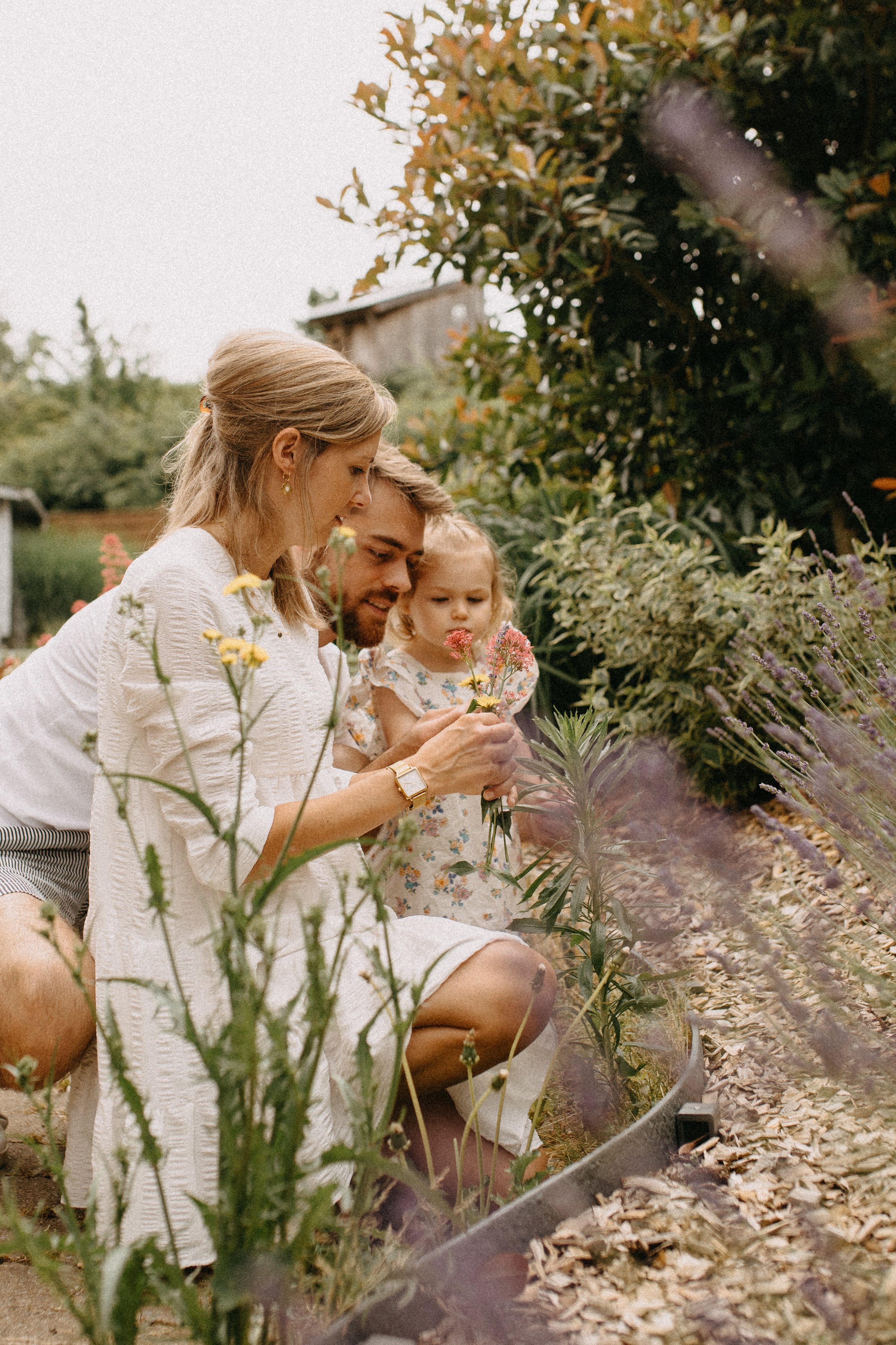 Une famille de trois personnes, dont une femme, un homme et une petite fille, examine des fleurs dans un jardin. La scène se déroule en plein air, avec des plantes et des arbustes en arrière-plan.
