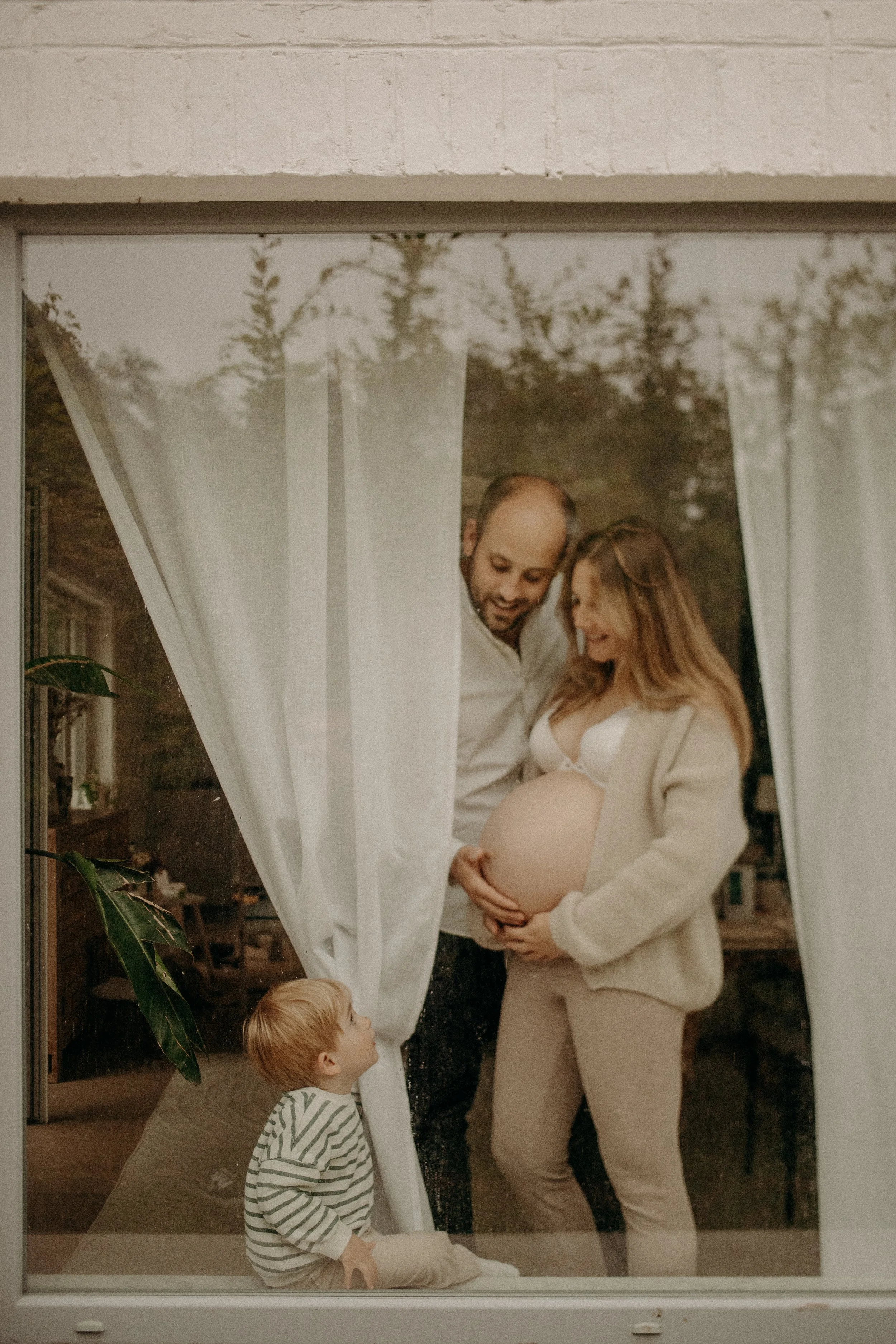 Une famille regarde à travers une fenêtre. La femme enceinte, le mari et un enfant regardent vers elle. La famille est à l'intérieur d'une maison.