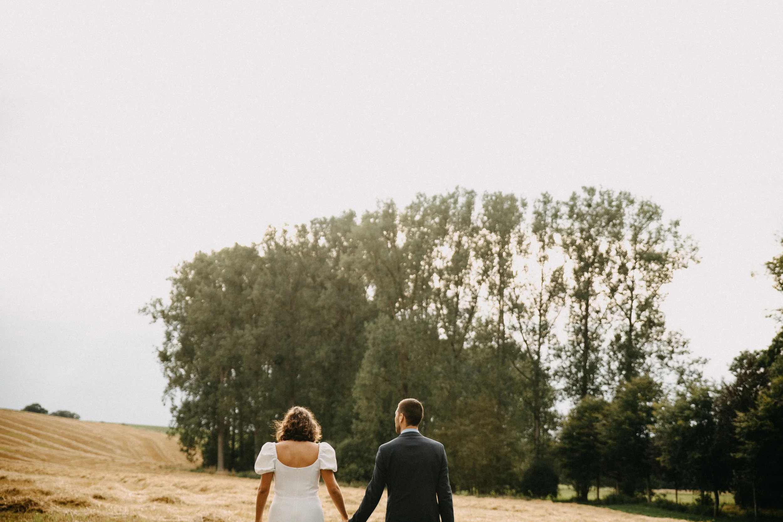 Un couple habillé formellement, marchant main dans la main dans un champ rural avec des arbres en arrière-plan.