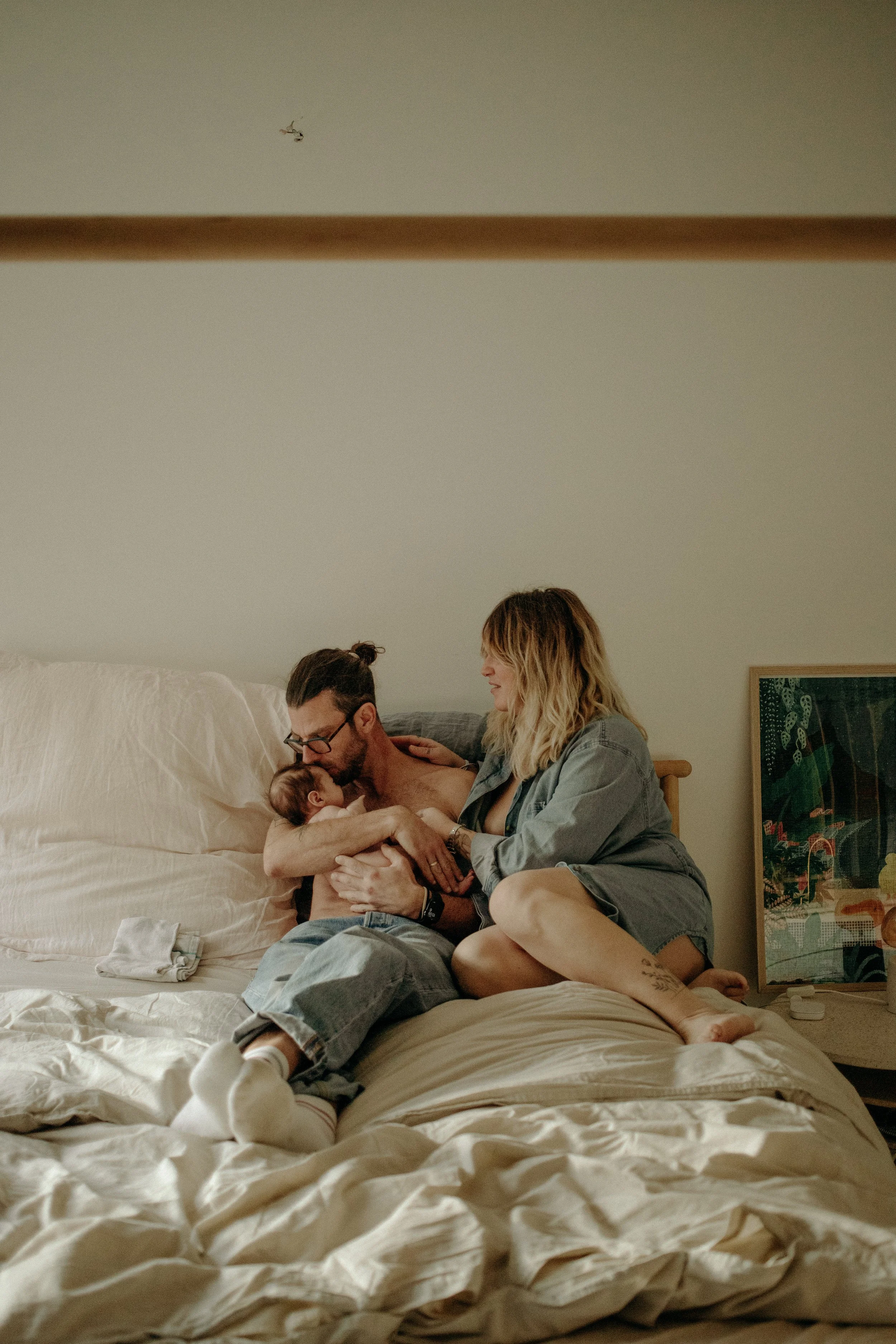 Une famille dans une chambre, un père, une mère et un bébé, partageant un moment intime sur un lit, ambiance chaleureuse et décontractée.