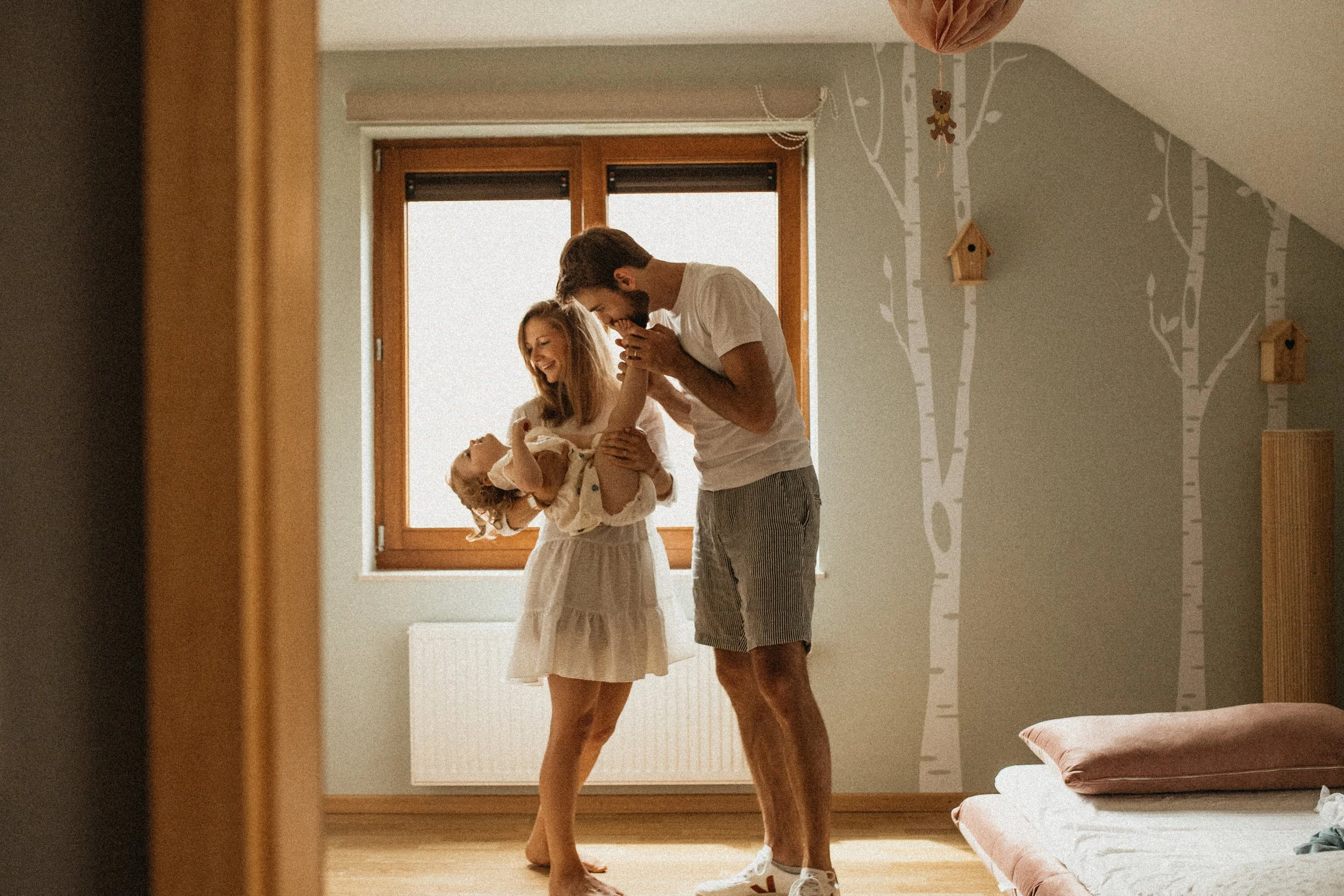 Une famille souriante, composée d'une mère, d'un père et d'une fille, partageant un moment de joie dans une chambre lumineuse avec un décor de forêt sur le mur.