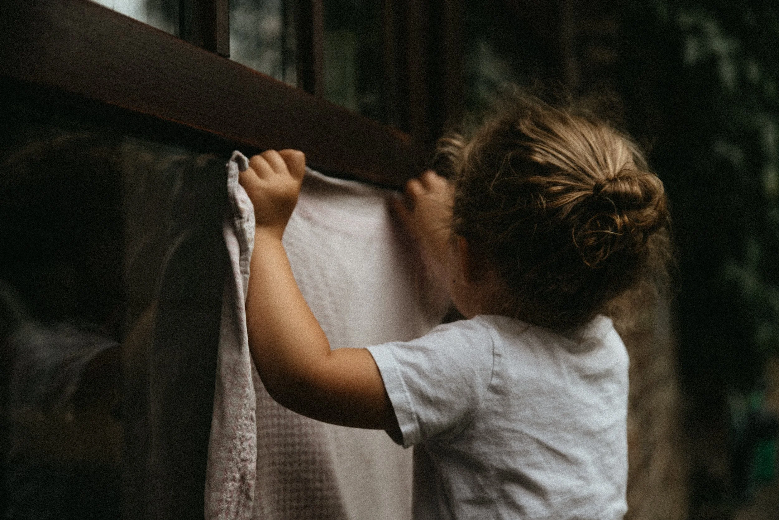 Un enfant touche une fenêtre à travers un rideau léger, dans une atmosphère chaleureuse et sombre.