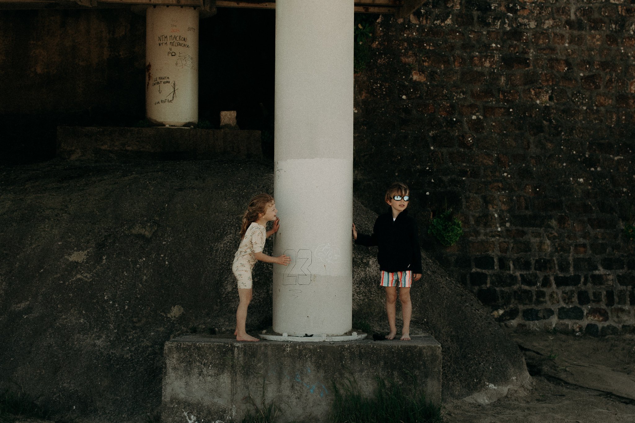 Deux enfants jouent derrière un poteau blanc dans une zone urbaine, un garçon portant des lunettes de soleil et un garçon en pyjama rayé et un haut noir, sous un pont avec des murs en briques.