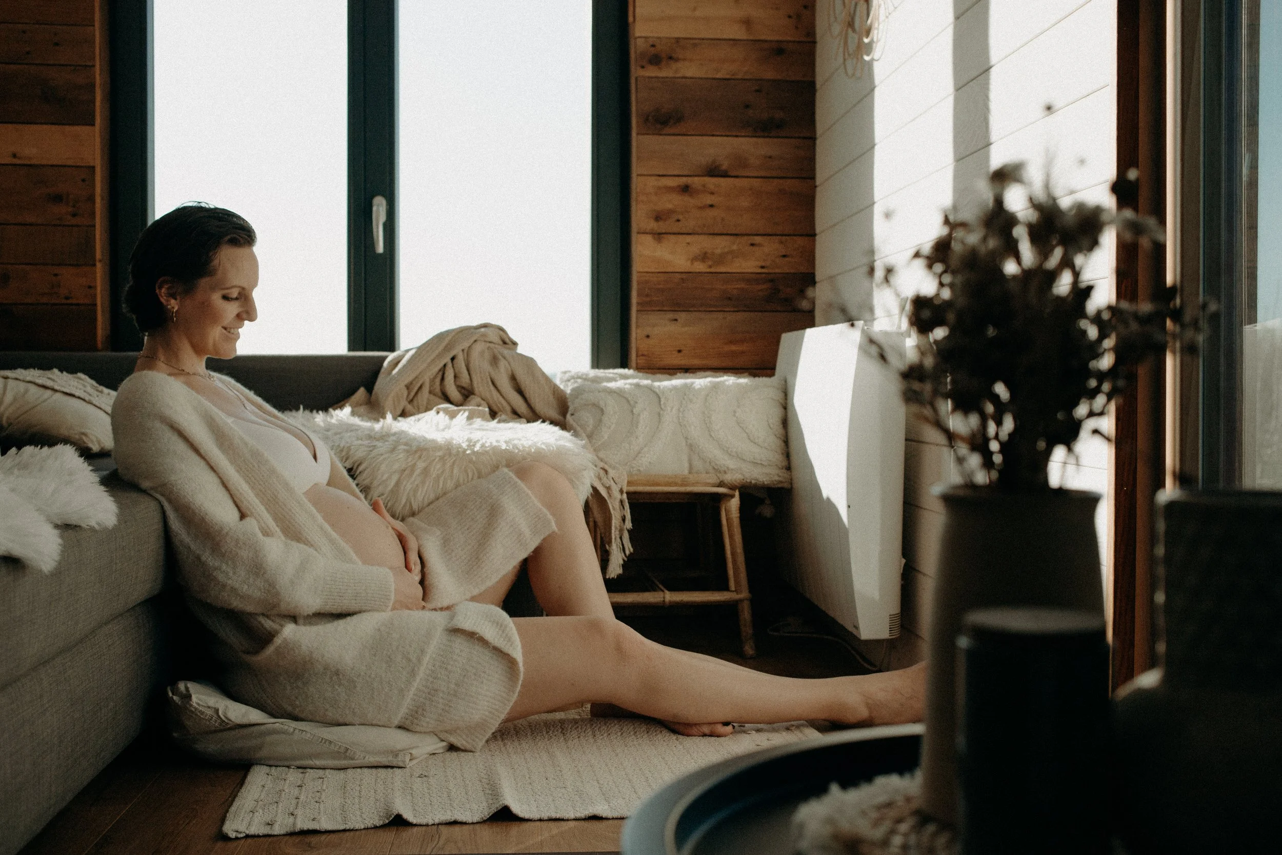 Une femme enceinte assise sur un sol dans un salon en bois, souriant, avec un grand miroir derrière elle, entourée de coussins et de vêtements confortables, dans une pièce lumineuse.