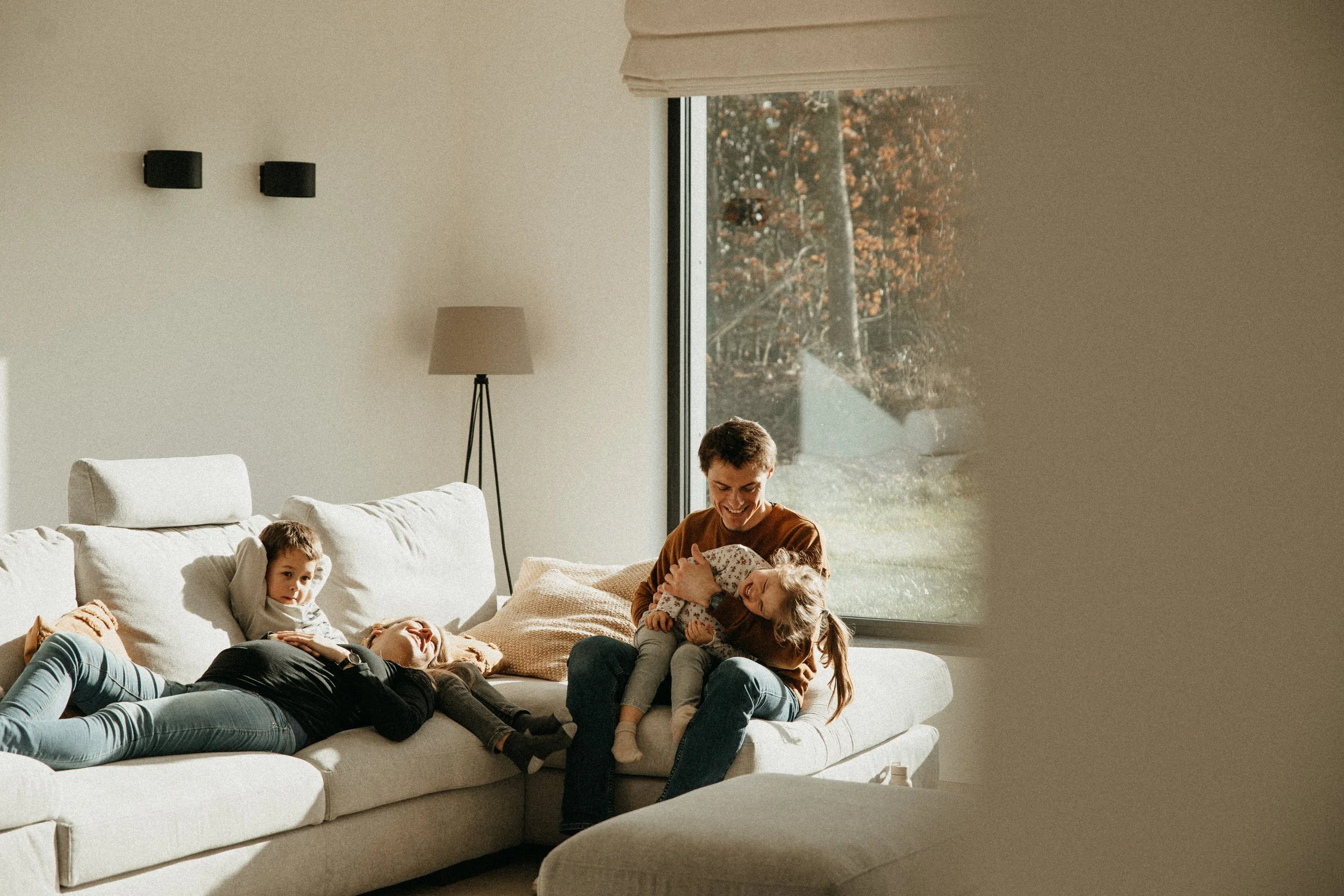 Une famille souriante jouant sur un canapé dans un salon lumineux avec un grand fenêtre et une vue sur le jardin en automne.