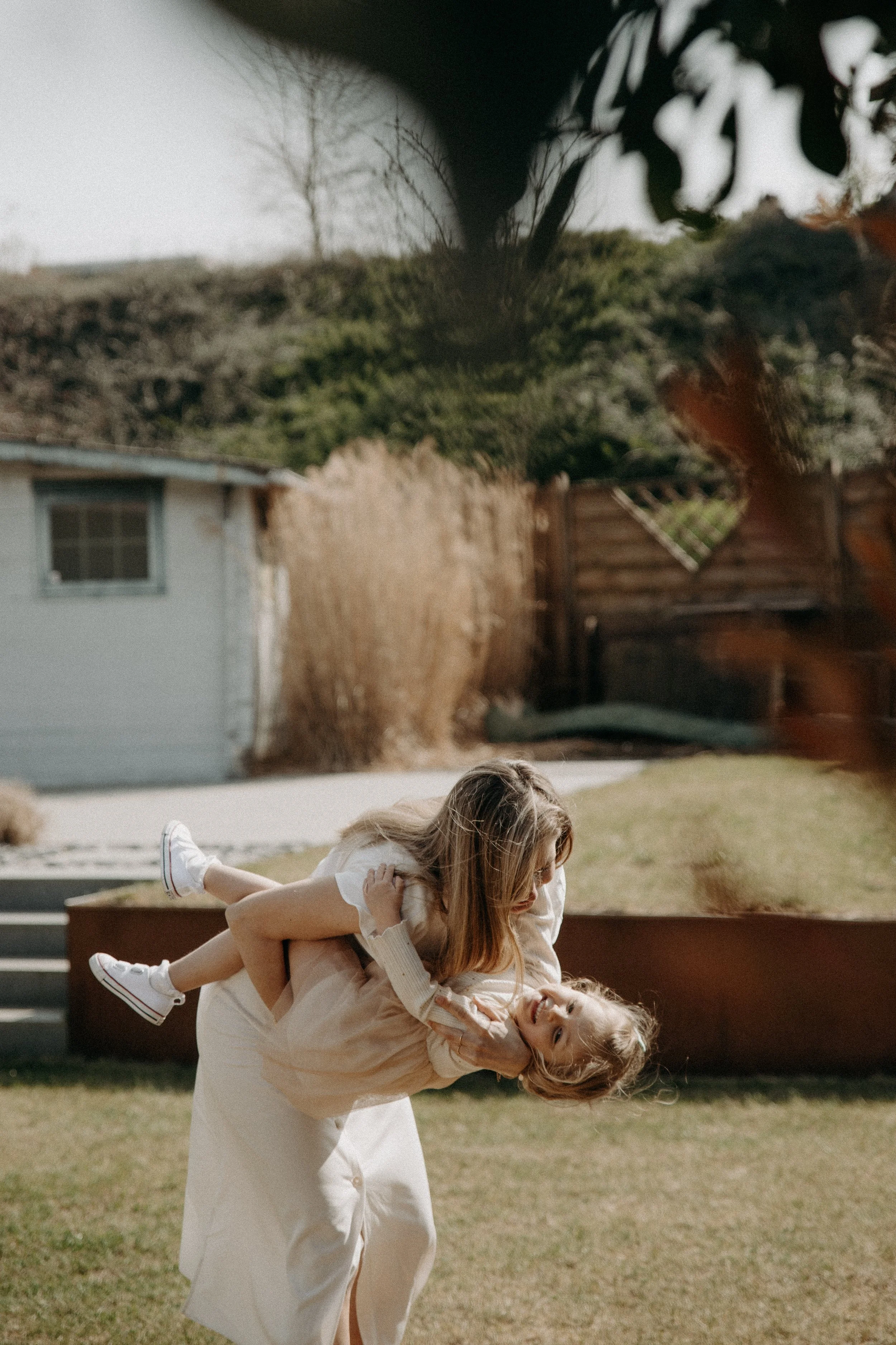 Deux femmes jouent dans un jardin en plein air, avec une maison en arrière-plan. L'une d'elles porte une robe blanche, l'autre un haut beige. Elles semblent s'amuser ensemble.