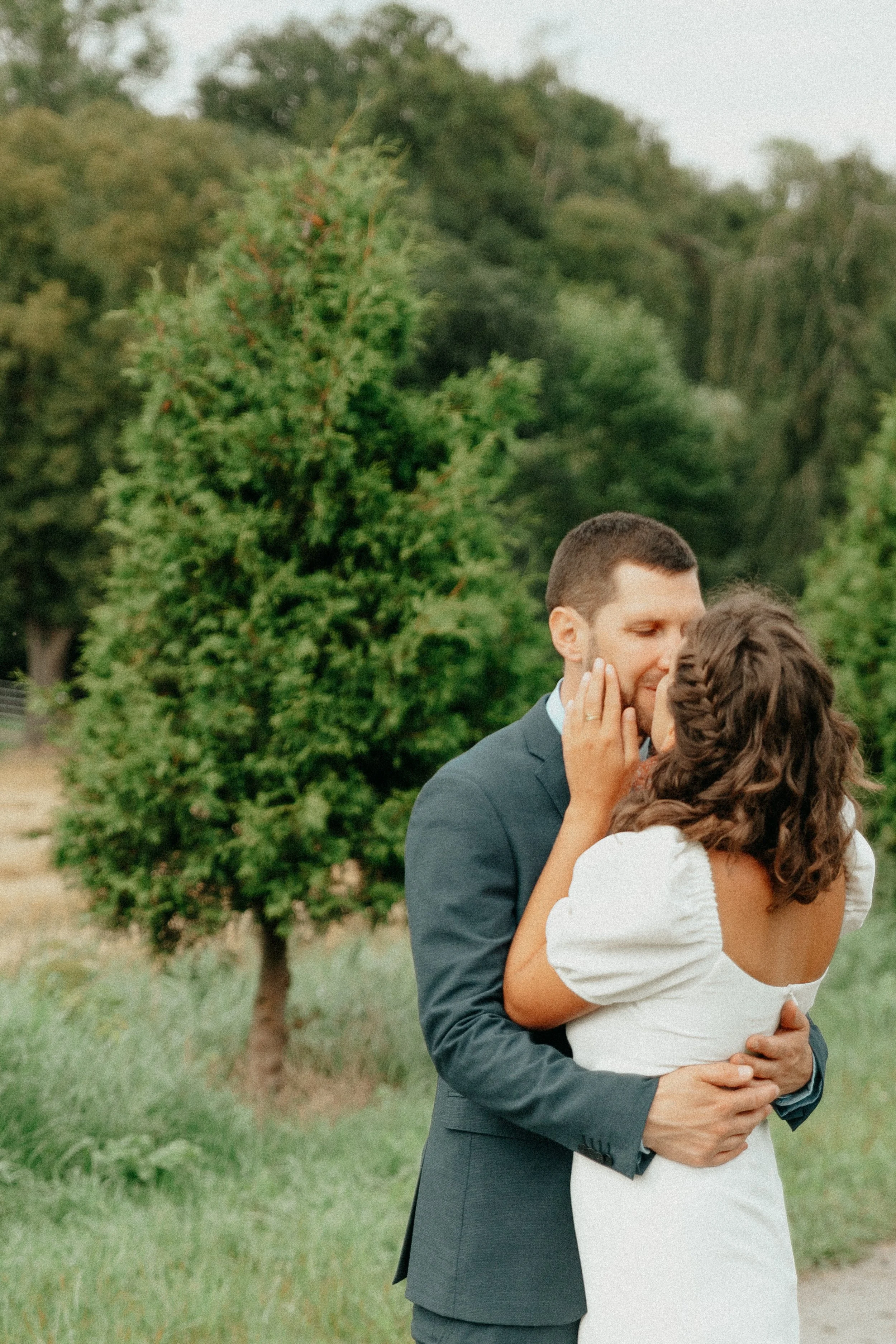 Un couple s'embrasse dans un parc, le homme porte un costume et la femme une robe blanche, avec des arbres en arrière-plan.