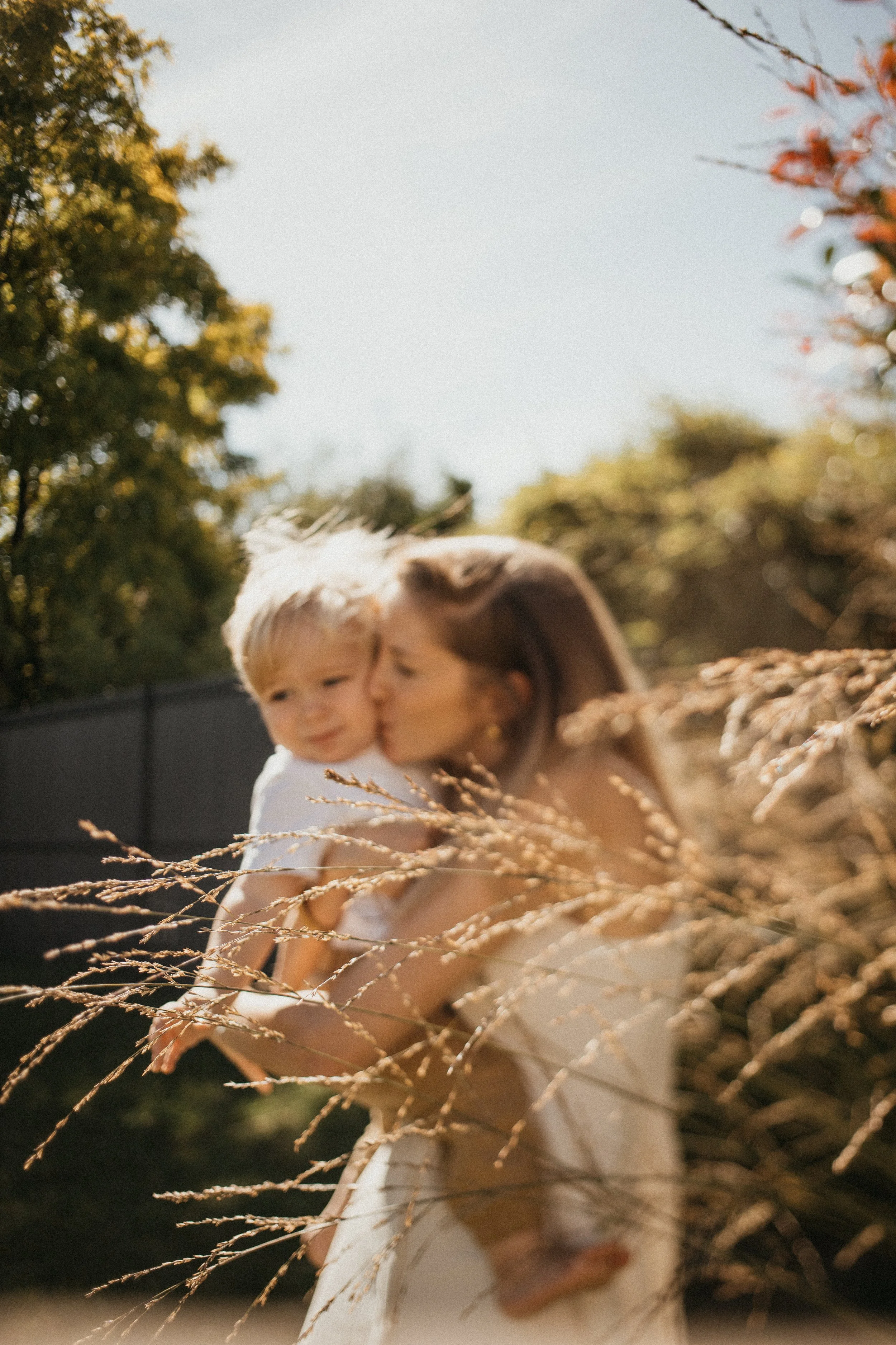 Une femme tient un petit garçon dans ses bras, elle lui donne un baiser sur la joue dans un jardin ensoleillé avec des arbres et des plantes.