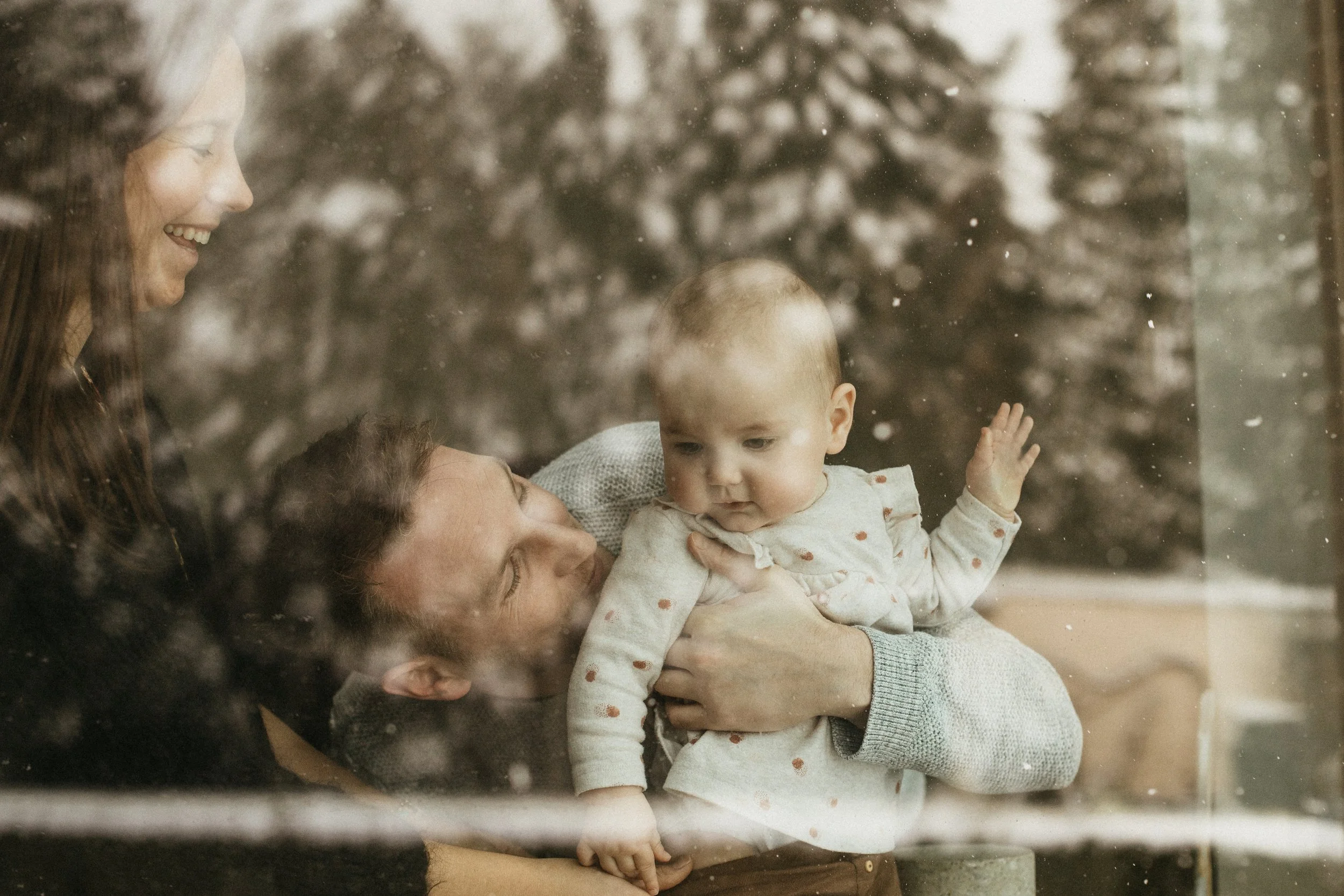 Une famille regarde par une vitre en hiver, avec un arbre enneigé en arrière-plan. Un homme tient un bébé et une femme sourit en les regardant.