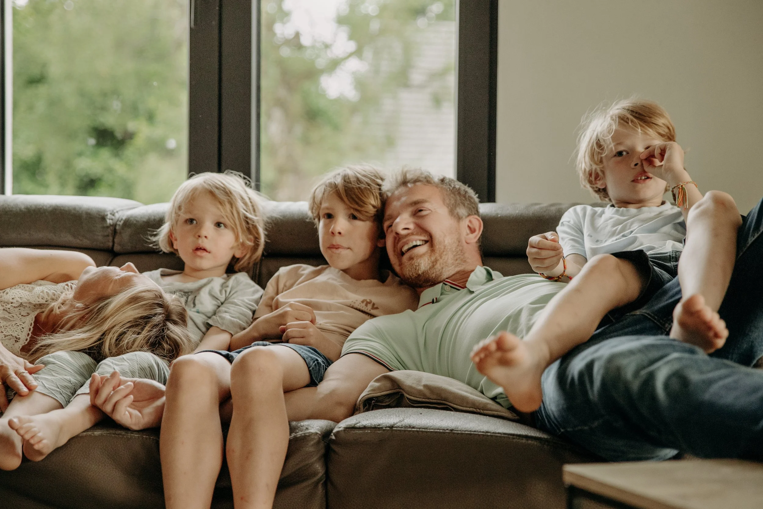 Une famille de six personnes, composée d'un adulte et de cinq enfants, qui se détend et rigole sur un canapé en intérieur avec une grande fenêtre derrière eux laissant entrer la lumière naturelle.