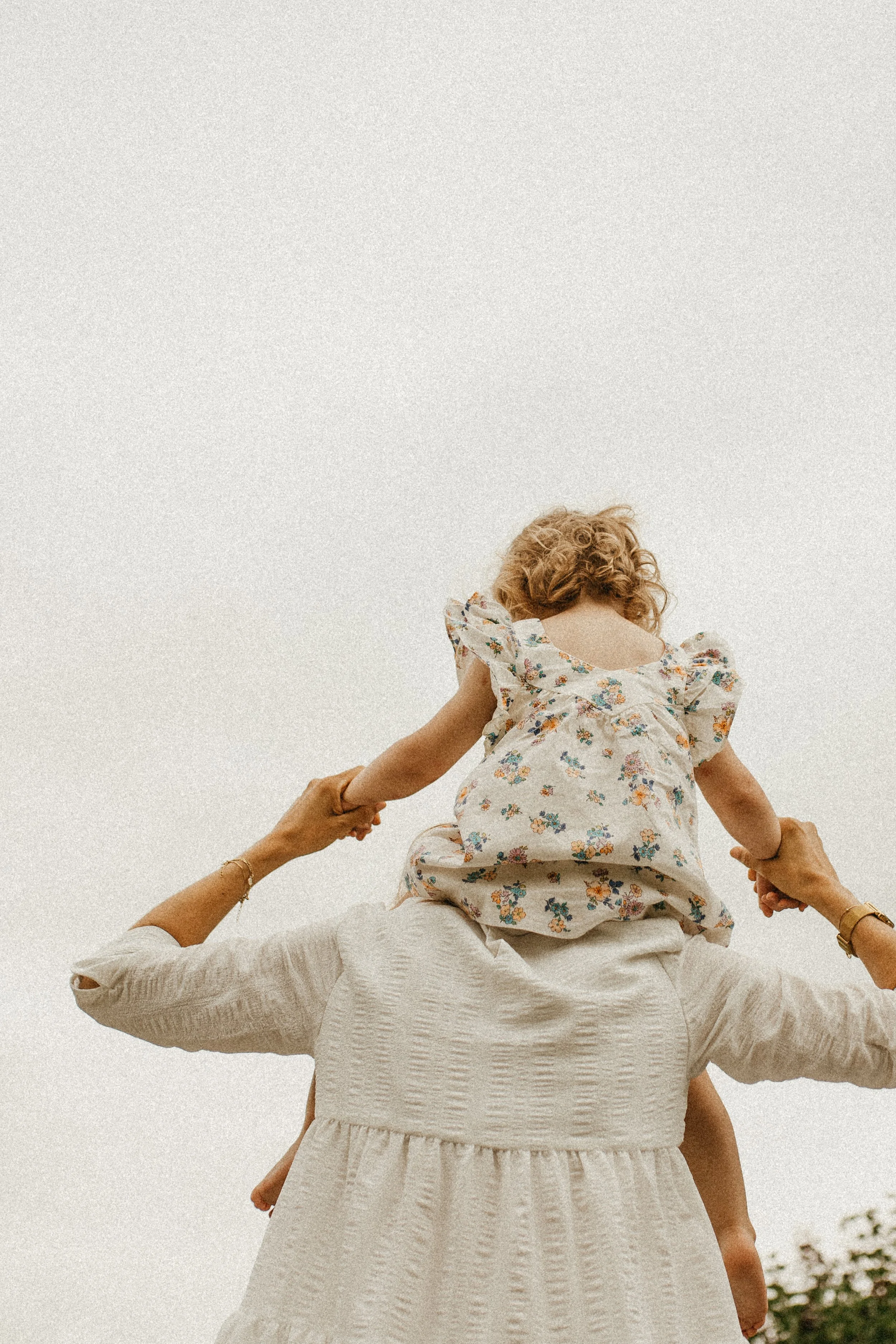 Une femme porte une jeune fille sur ses épaules, toutes deux tiennent la main, contre un ciel clair.