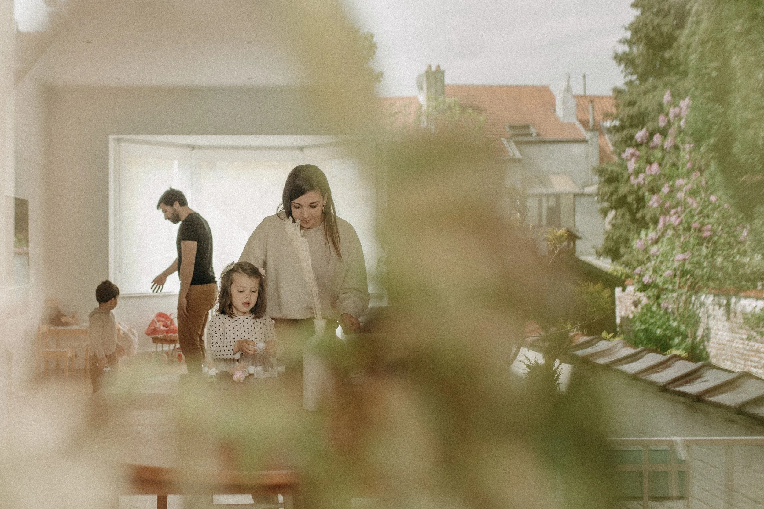 Une famille composée de deux adultes et deux enfants joue ensemble dans une pièce lumineuse avec des fenêtres, le tout vu partiellement à travers des feuilles floues à l'avant.