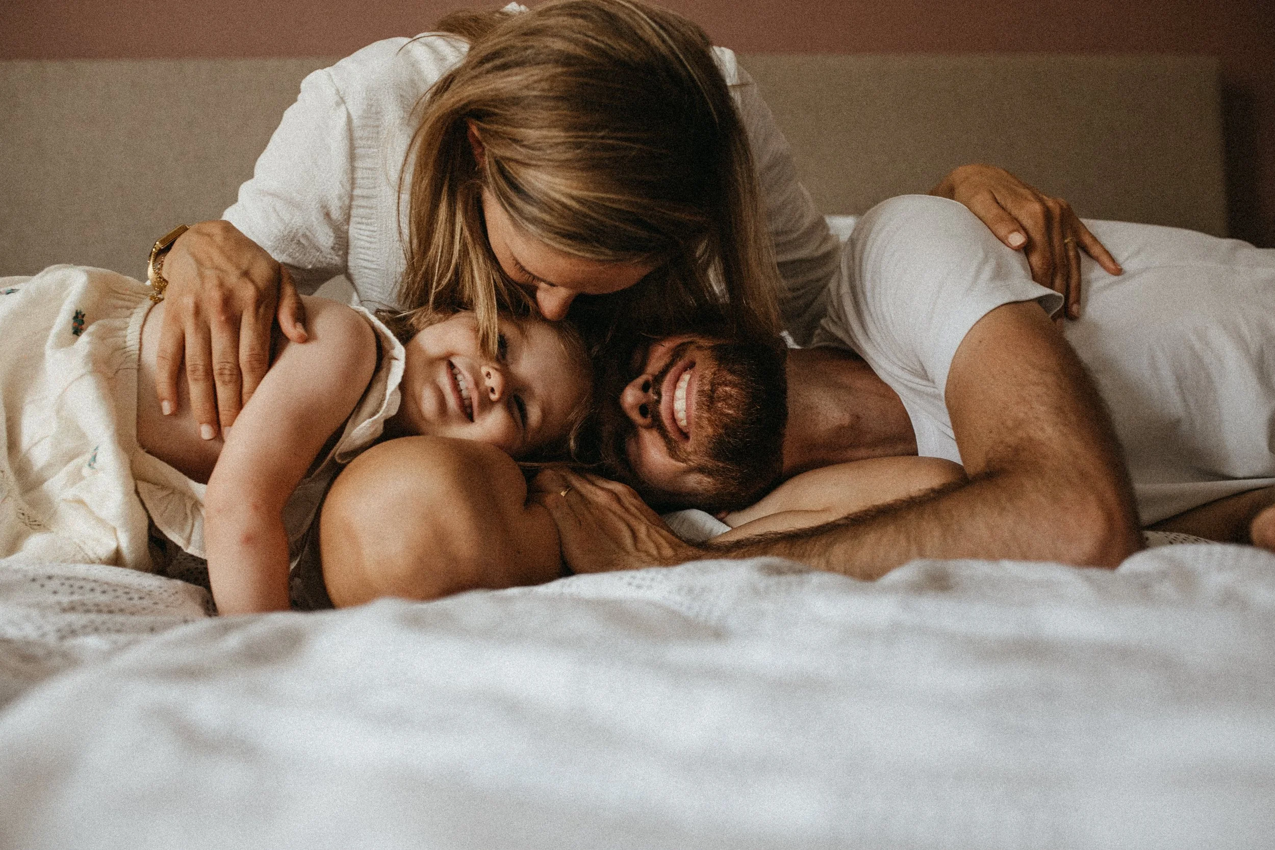 Une famille de trois personnes, un homme, une femme et un jeune garçon, partage un moment de tendresse et de joie dans un lit, avec des expressions de bonheur.