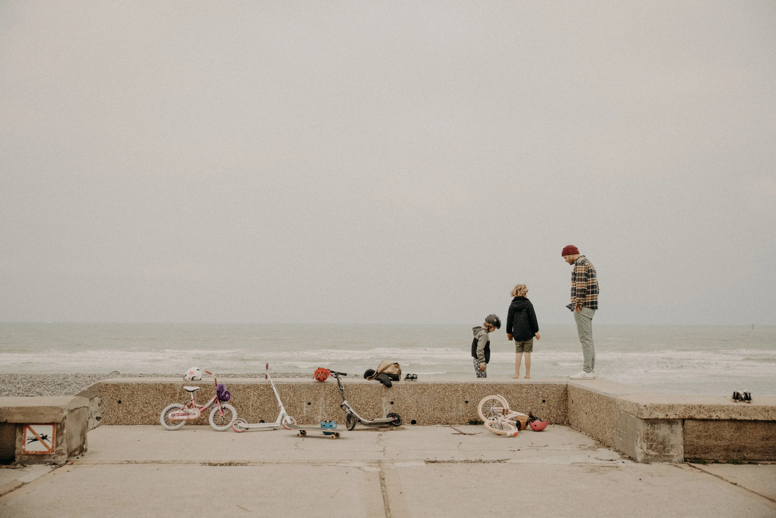 Une famille à la plage avec des vélos et des scooters, regardant la mer dans un ciel nuageux.