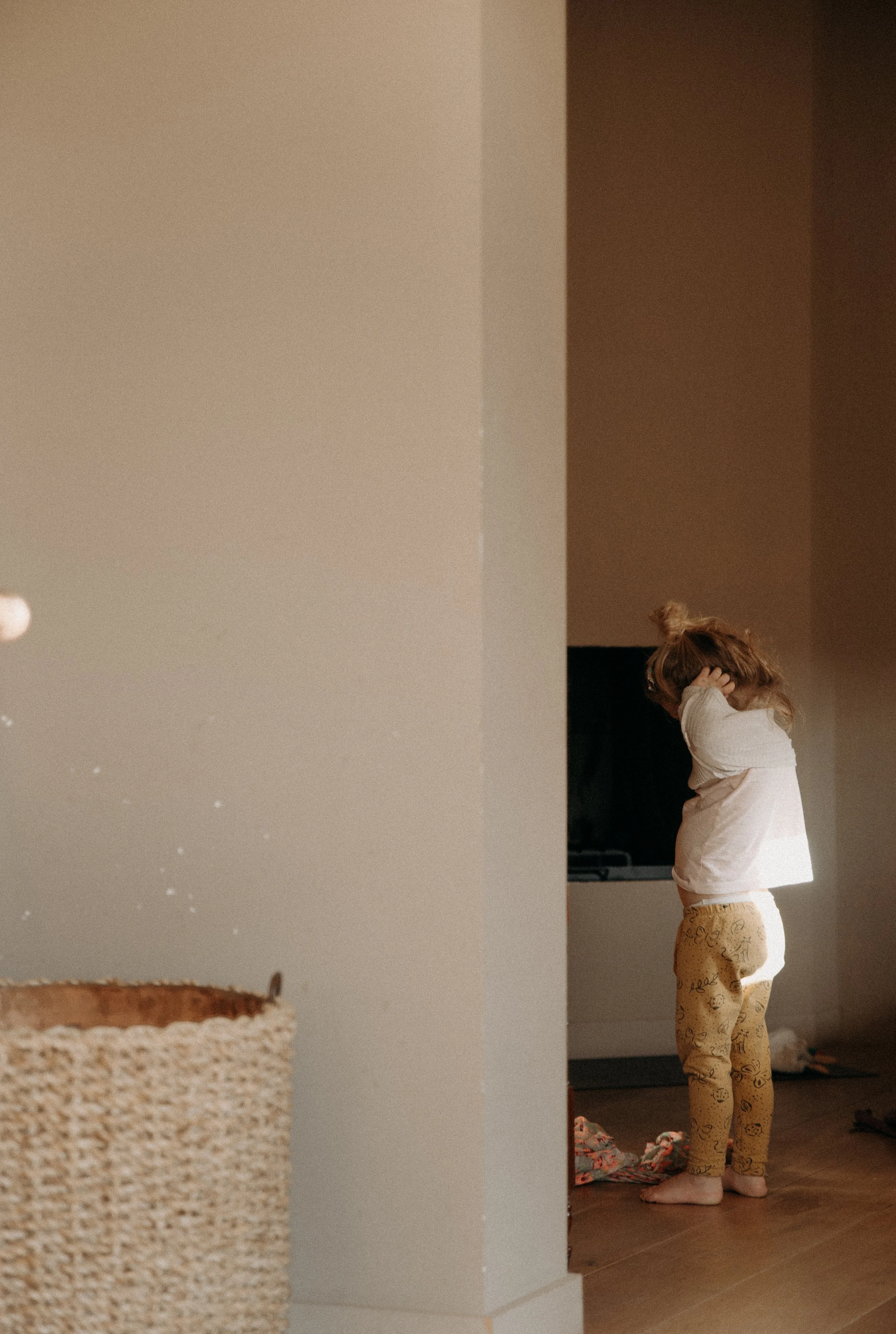 Une petite fille se tient dans un coin de la pièce, ajustant ses cheveux ou son bonnet, vêtue d’un pyjama jaune avec des dessins, dans une ambiance chaleureuse et tamisée.