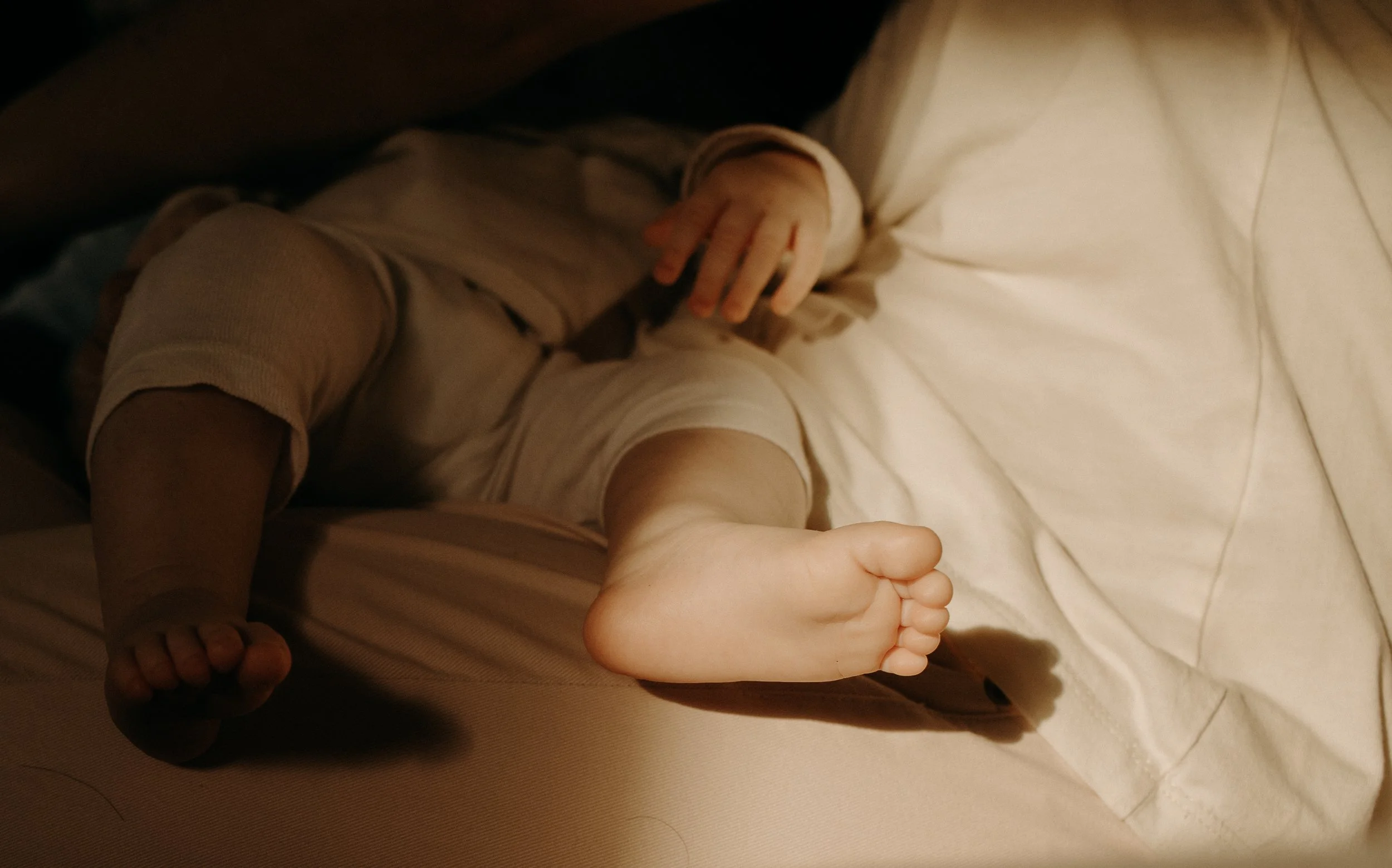 Un bébé allongé sur un lit, vêtu d'un pyjama beige, avec un bras tombant sur la surface du lit et la jambe en l'air, vu de côté.