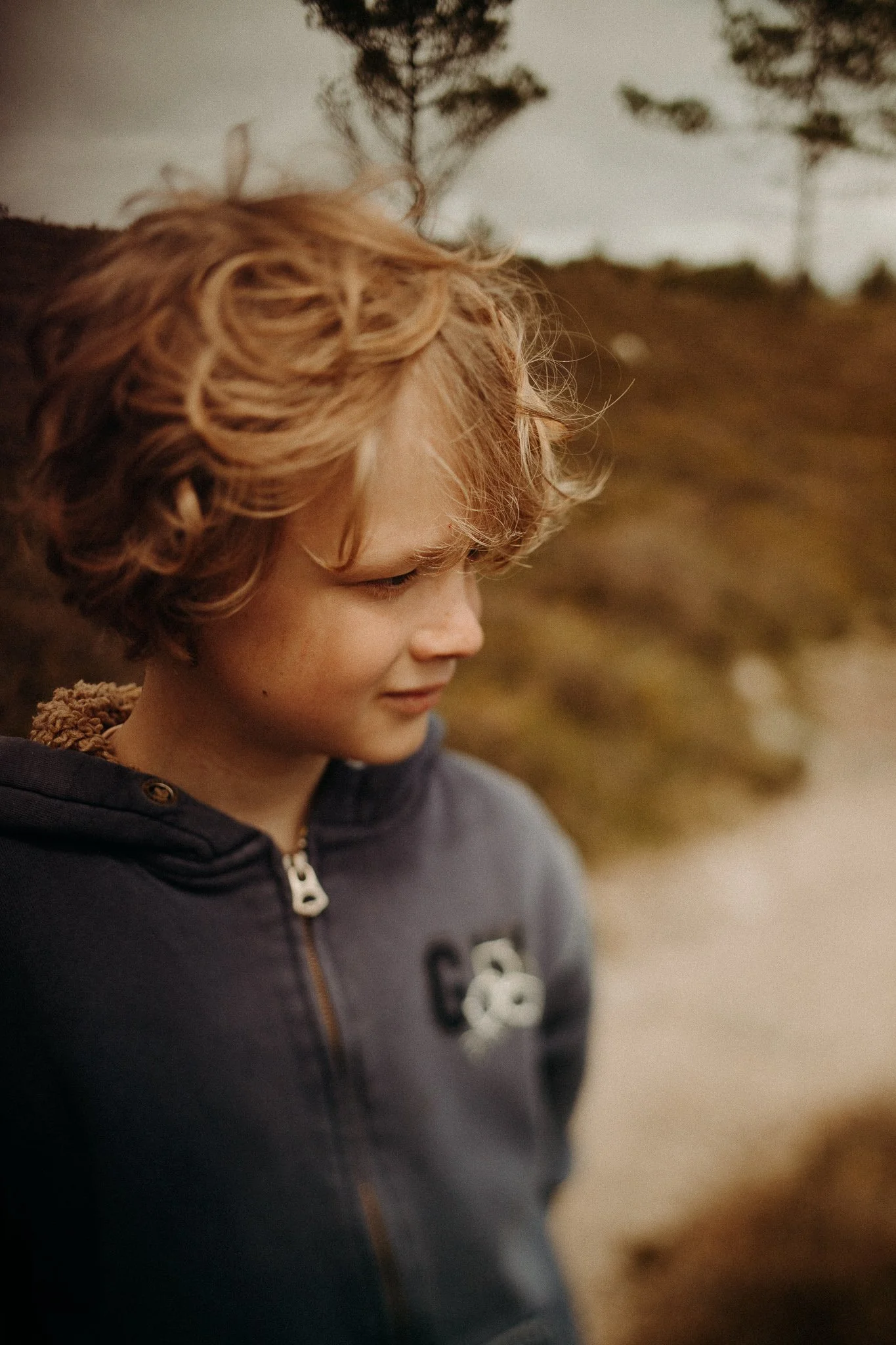 Jeune garçon aux cheveux bouclés, portant un sweat à capuche, regardant vers le bas, en extérieur avec des arbres en arrière-plan.