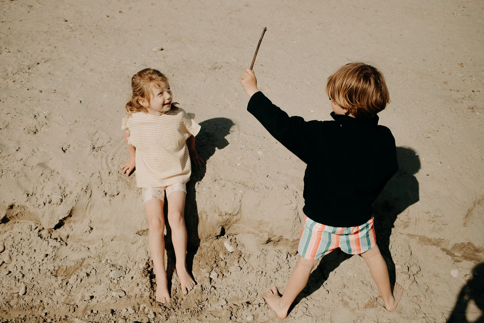 Deux enfants jouent sur la plage, un garçon avec un sweat noir et un short à rayures, une fille en chemise rayée, tous deux pieds nus sur le sable.