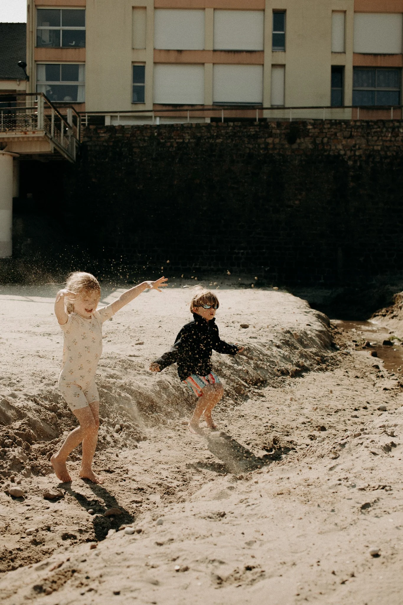 Deux enfants jouent et courent dans le sable d'une plage ensoleillée près d'une ville avec des bâtiments résidentiels en arrière-plan.