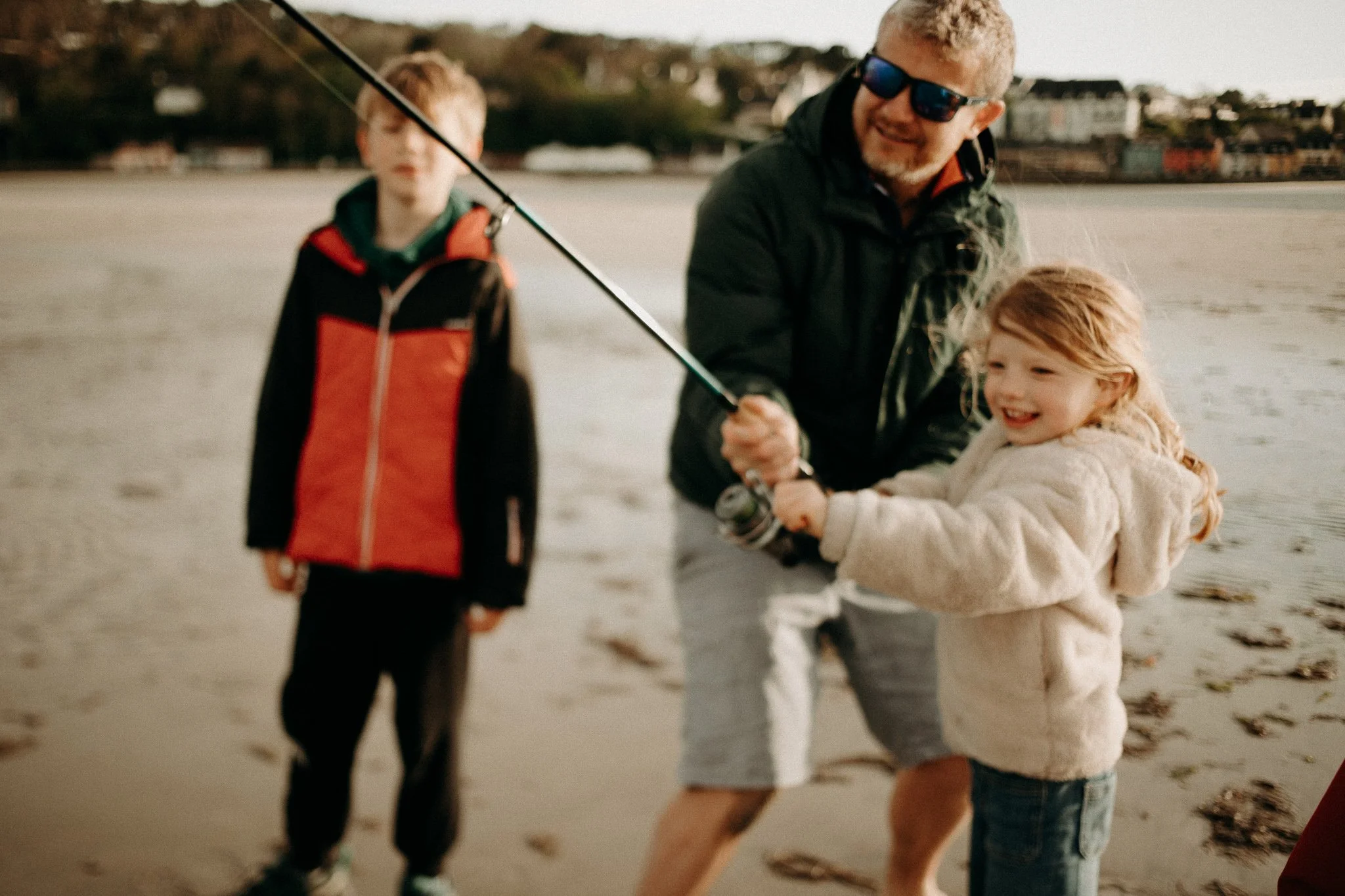 Un homme avec deux enfants, un garçon et une fille, sur une plage. Le père aide la fille à tenir une canne à pêche, et ils sourient tous les deux.