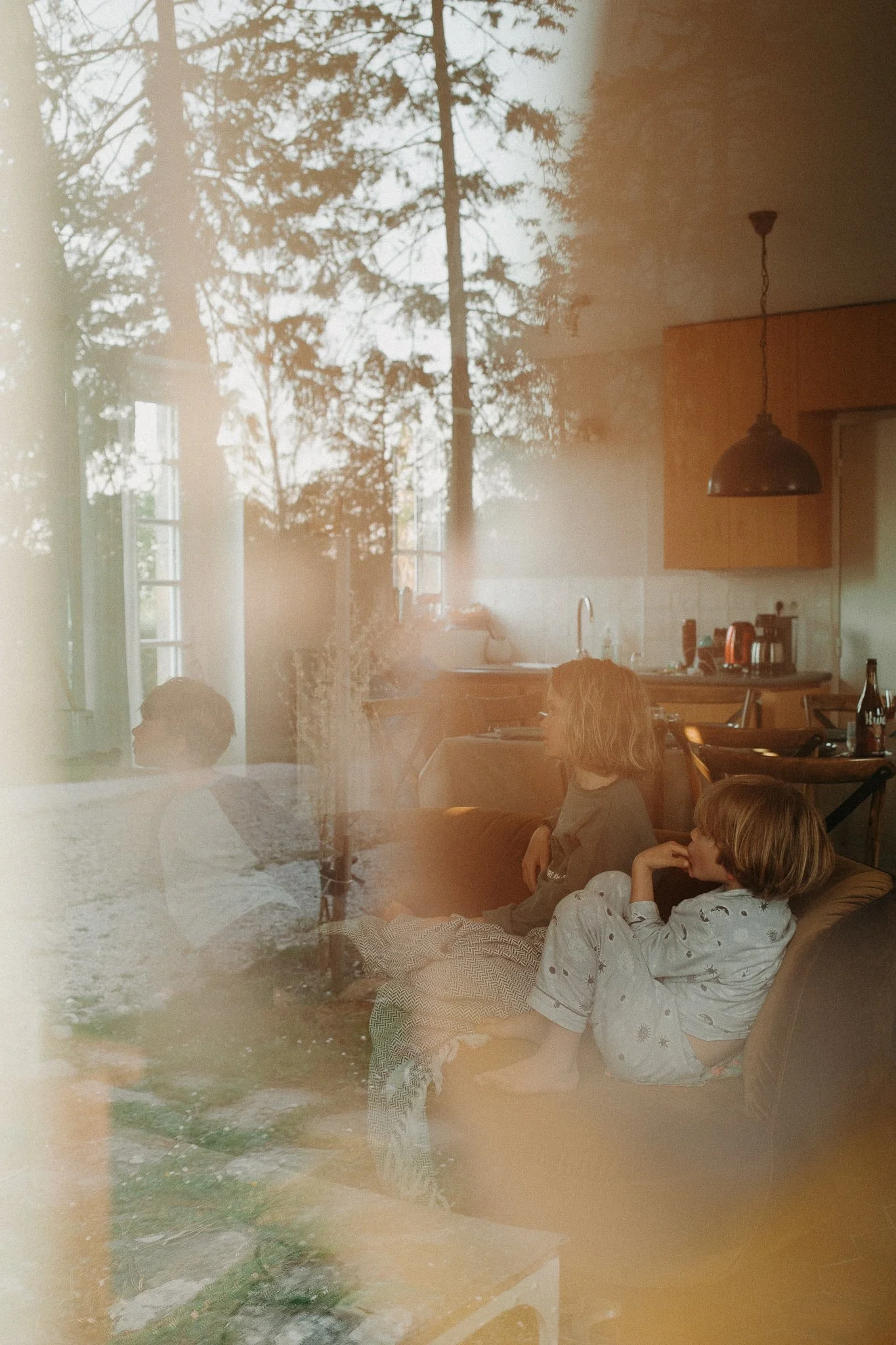 Une scène intérieure avec deux enfants assis sur un canapé, regardant vers la gauche. La pièce a une cuisine en arrière-plan, avec des fenêtres laissant entrer la lumière naturelle, et une double exposition avec une image extérieure d'arbres et de pi