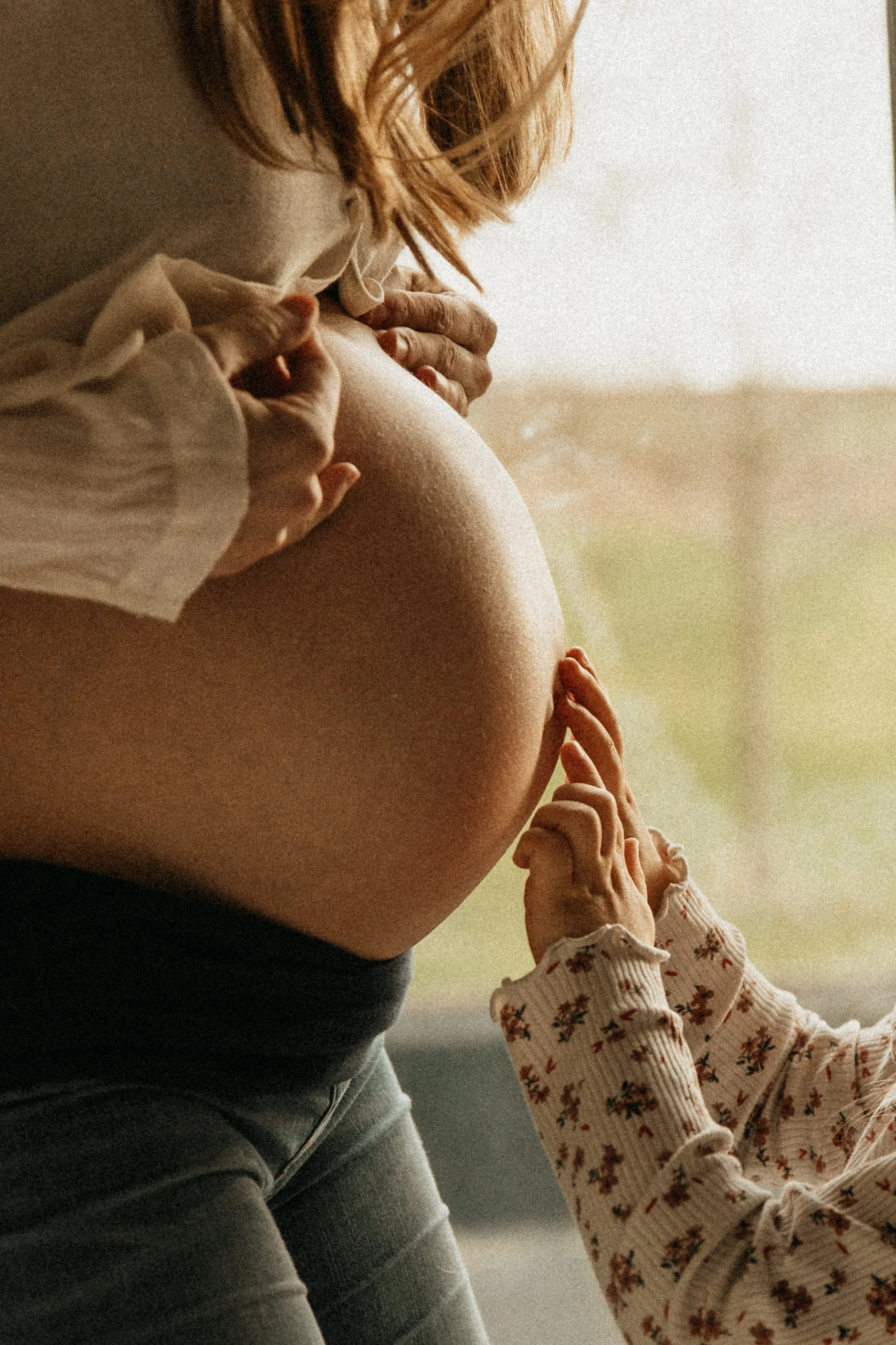 Une femme enceinte, vue de côté, avec une main soutenant son ventre rond, et une petite main d'enfant touchant son ventre, près de sa poitrine, dans un intérieur lumineux.