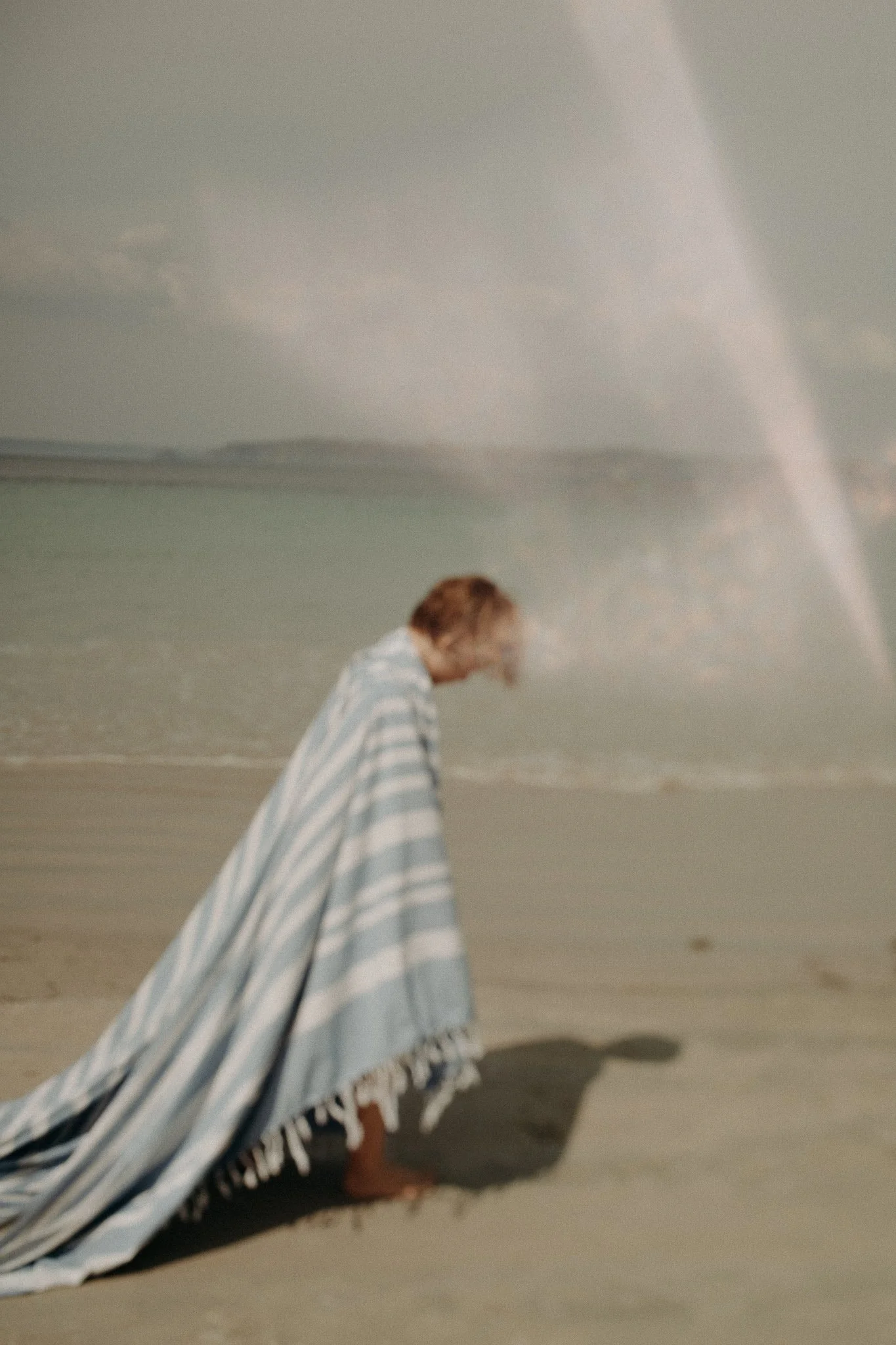 Une personne debout sur la plage enveloppée dans une couverture rayée, regardant l'océan.