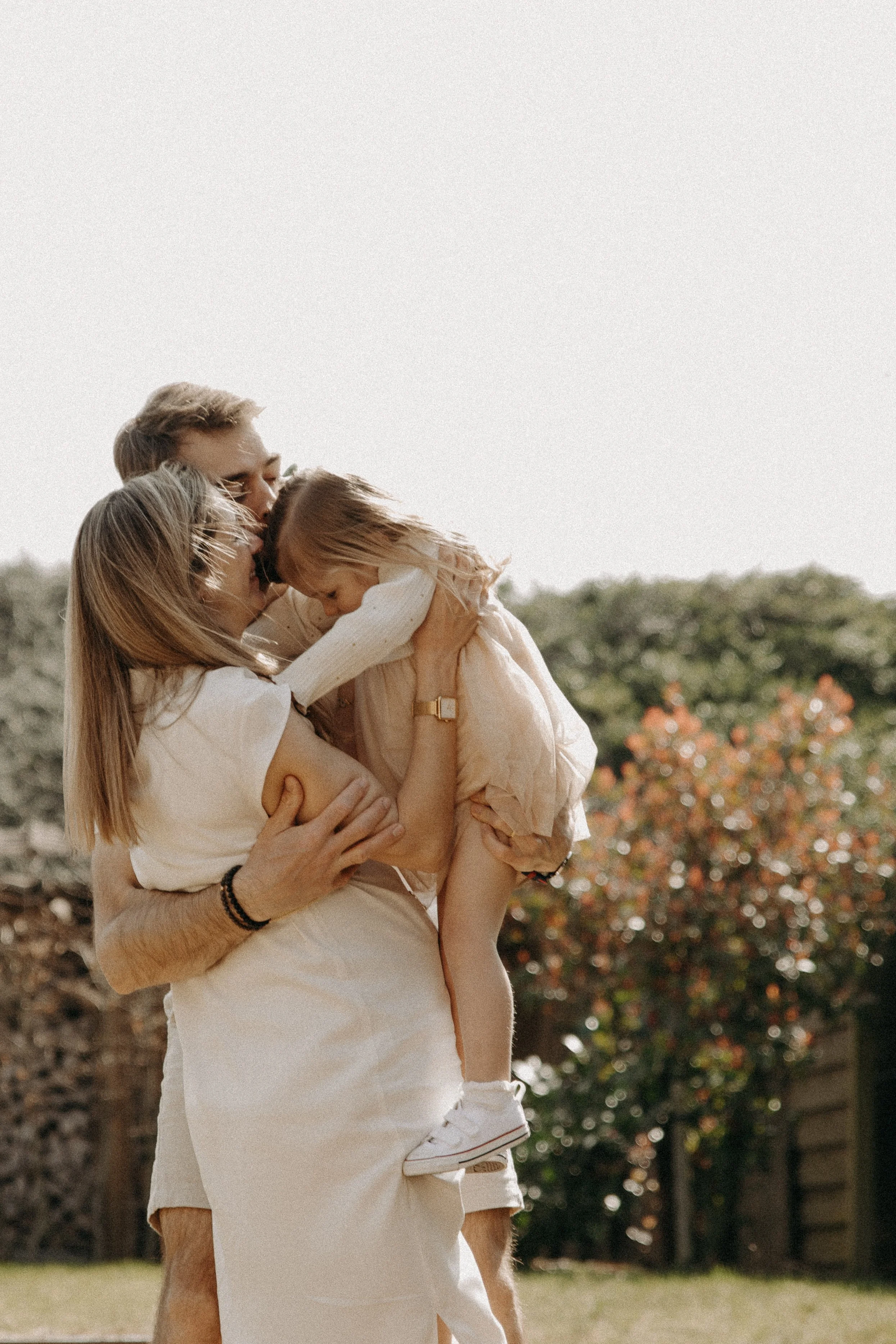 Une famille heureuse en extérieur, un père, une mère et leur fille blonde, s'embrassant et partageant un moment d'affection, sous un ciel clair avec des arbres en arrière-plan.