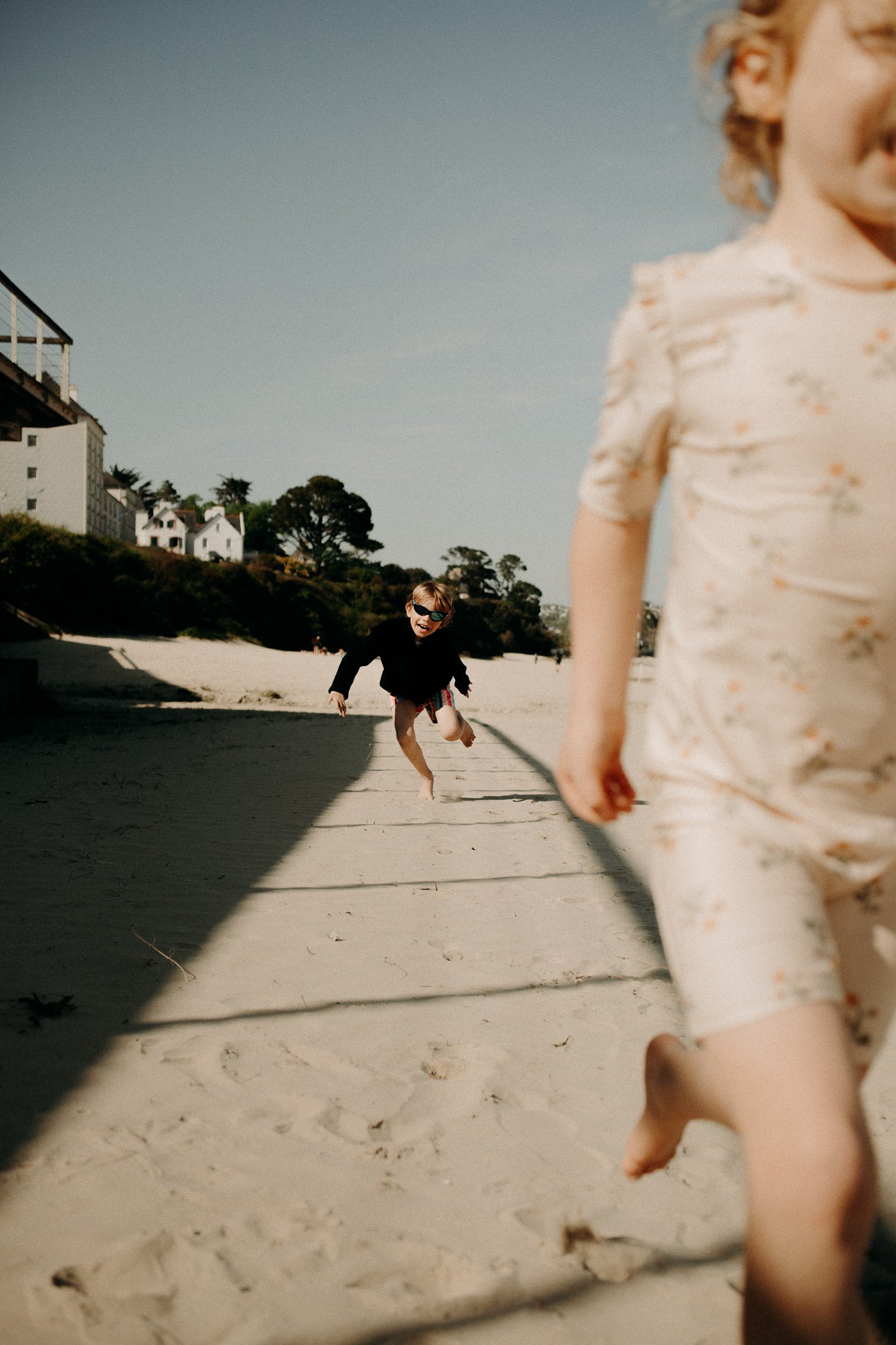 Deux enfants jouent et courent sur la plage ensoleillée, avec des maisons blanches et des arbres en arrière-plan.