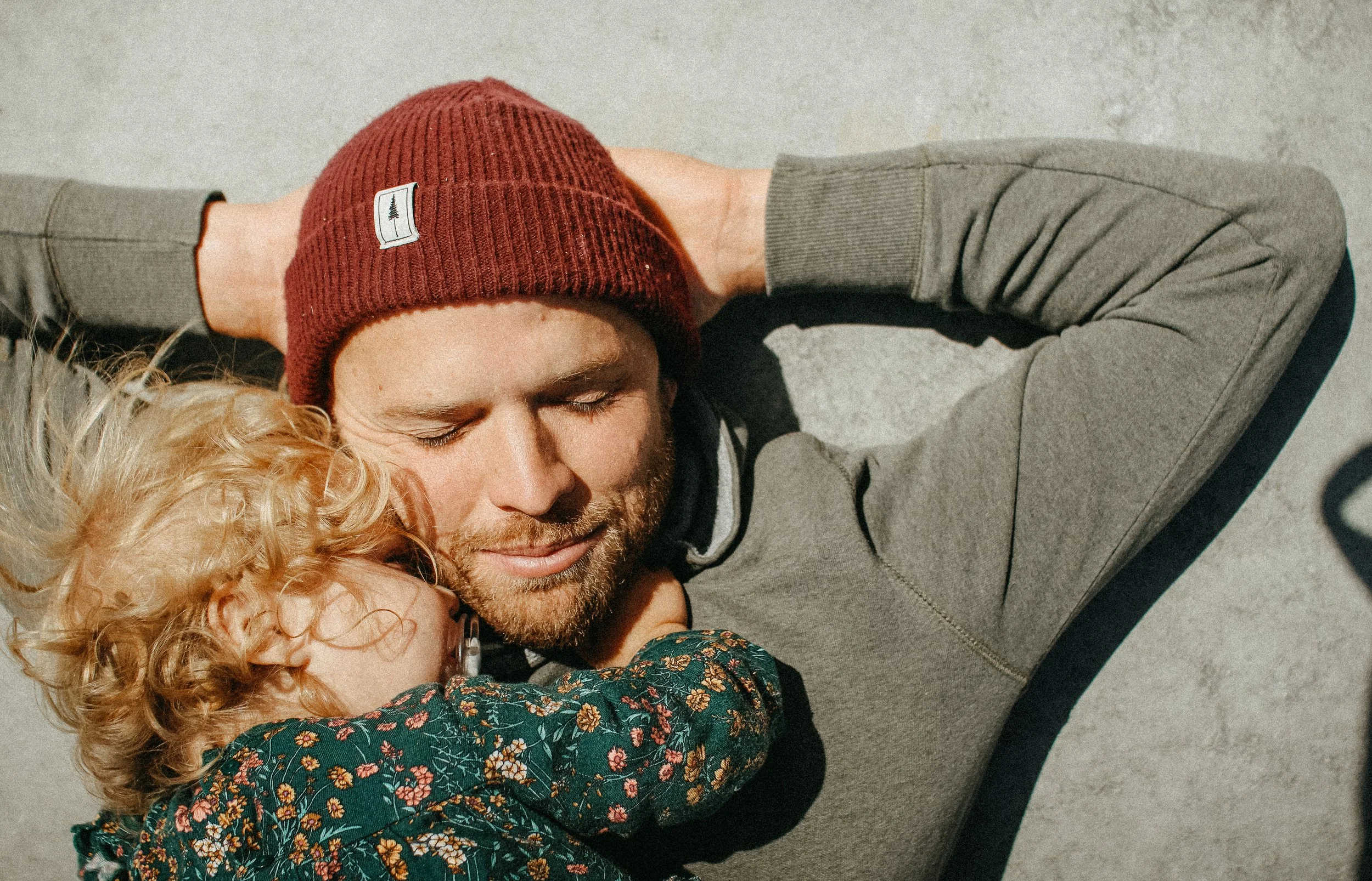Un homme et un enfant jouent et se câlinent sur le sol, l'homme porte un bonnet rouge et le petit a des cheveux bouclés et porte des lunettes et un vêtement à motif floral.
