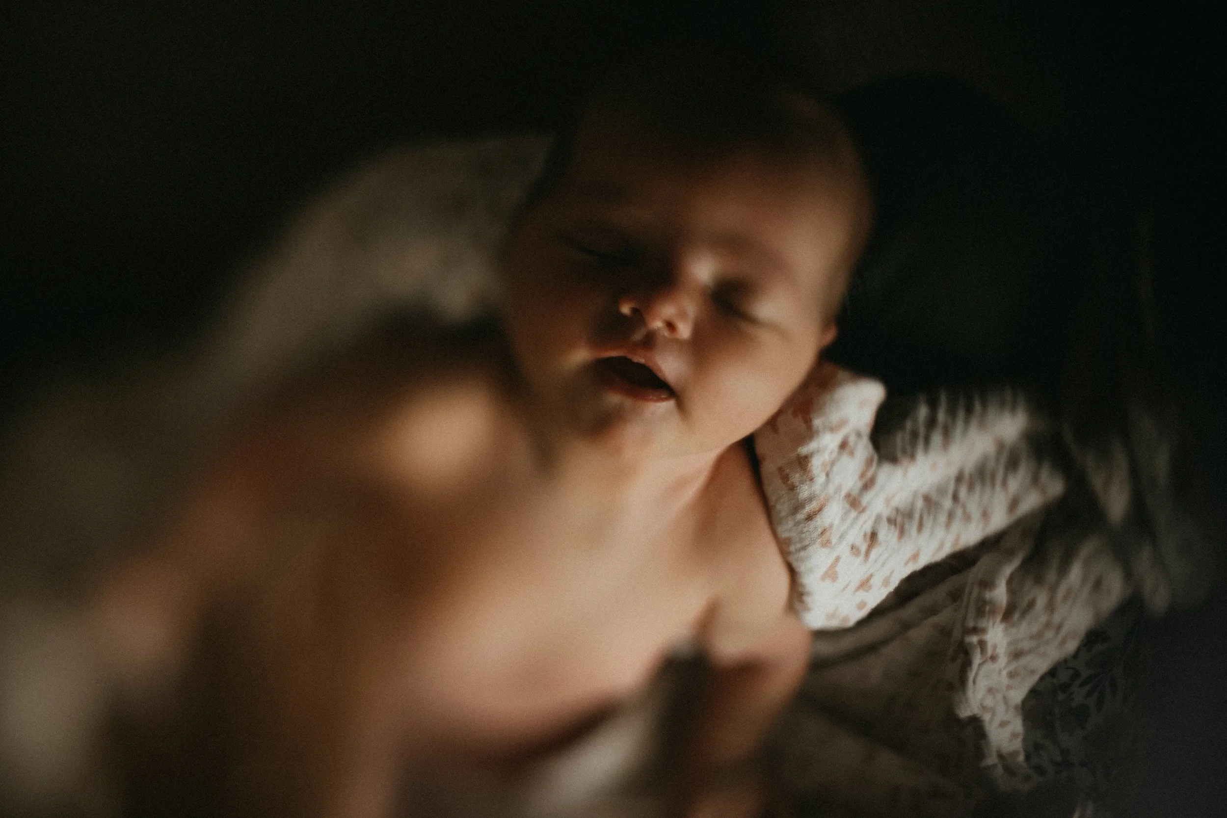 Un bébé endormi dans une couverture foncée, portant un vêtement à motifs beige et blanc.