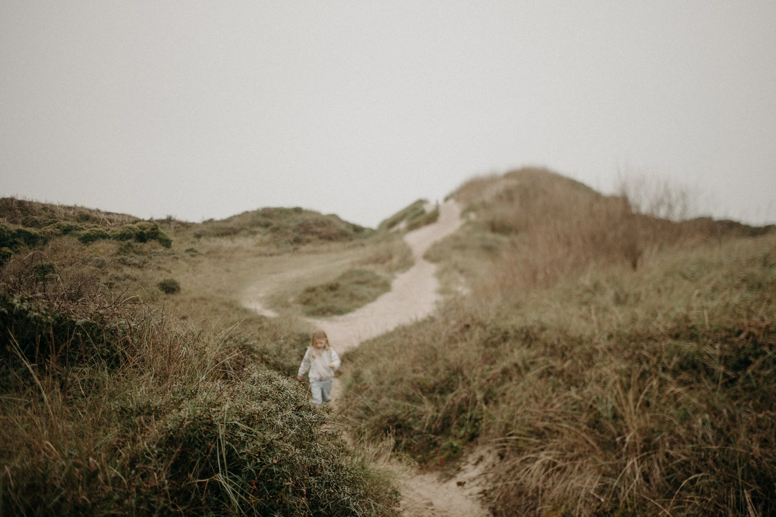 Une jeune fille marche sur un sentier dans un paysage de dunes et de végétation dense sous un ciel brumeux.