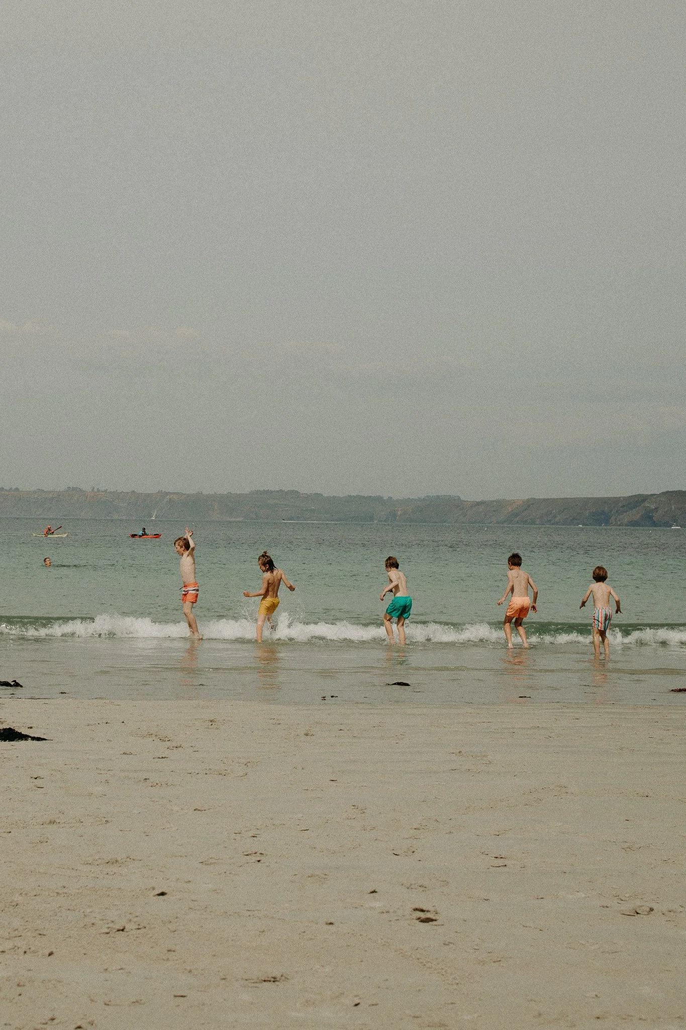 Enfants jouant et nageant dans la mer à la plage, avec des kayaks au loin dans l'eau et un ciel nuageux.