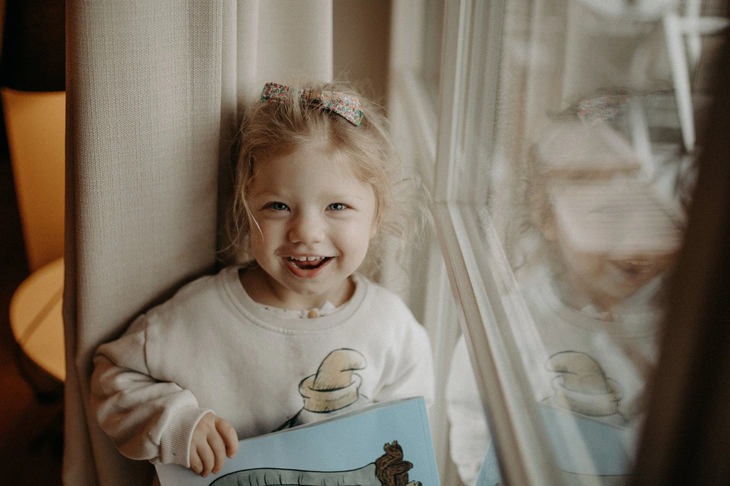 Une jeune fille blonde avec un ruban à cheveux coloré, souriante, tenant un livre illustré, regardant par la fenêtre, assise près d’un mur beige.