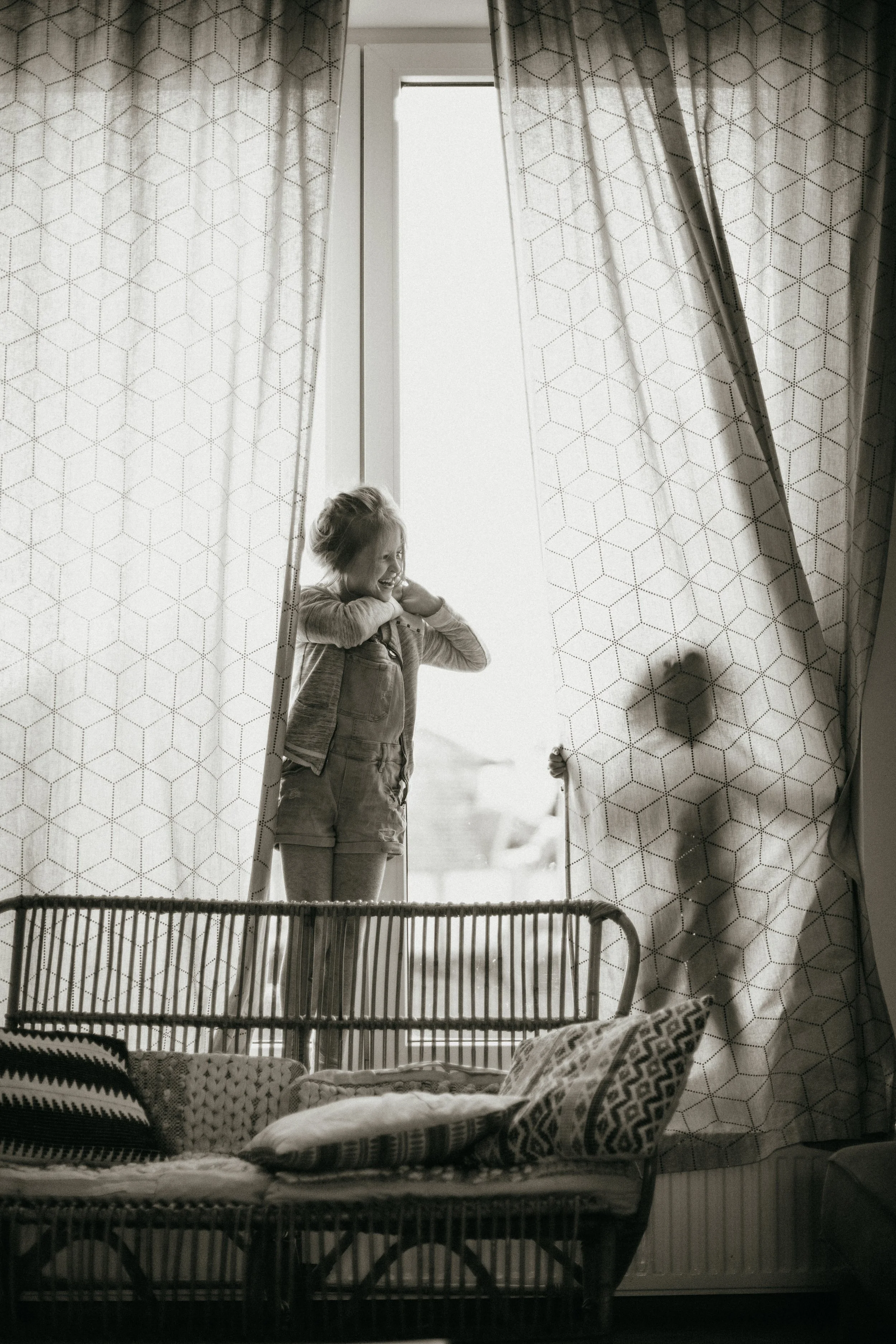 Une jeune fille regarde par une fenêtre en grande partie occultée par des rideaux, sa silhouette est visible dans la lumière, créant une ombre sur le rideau.