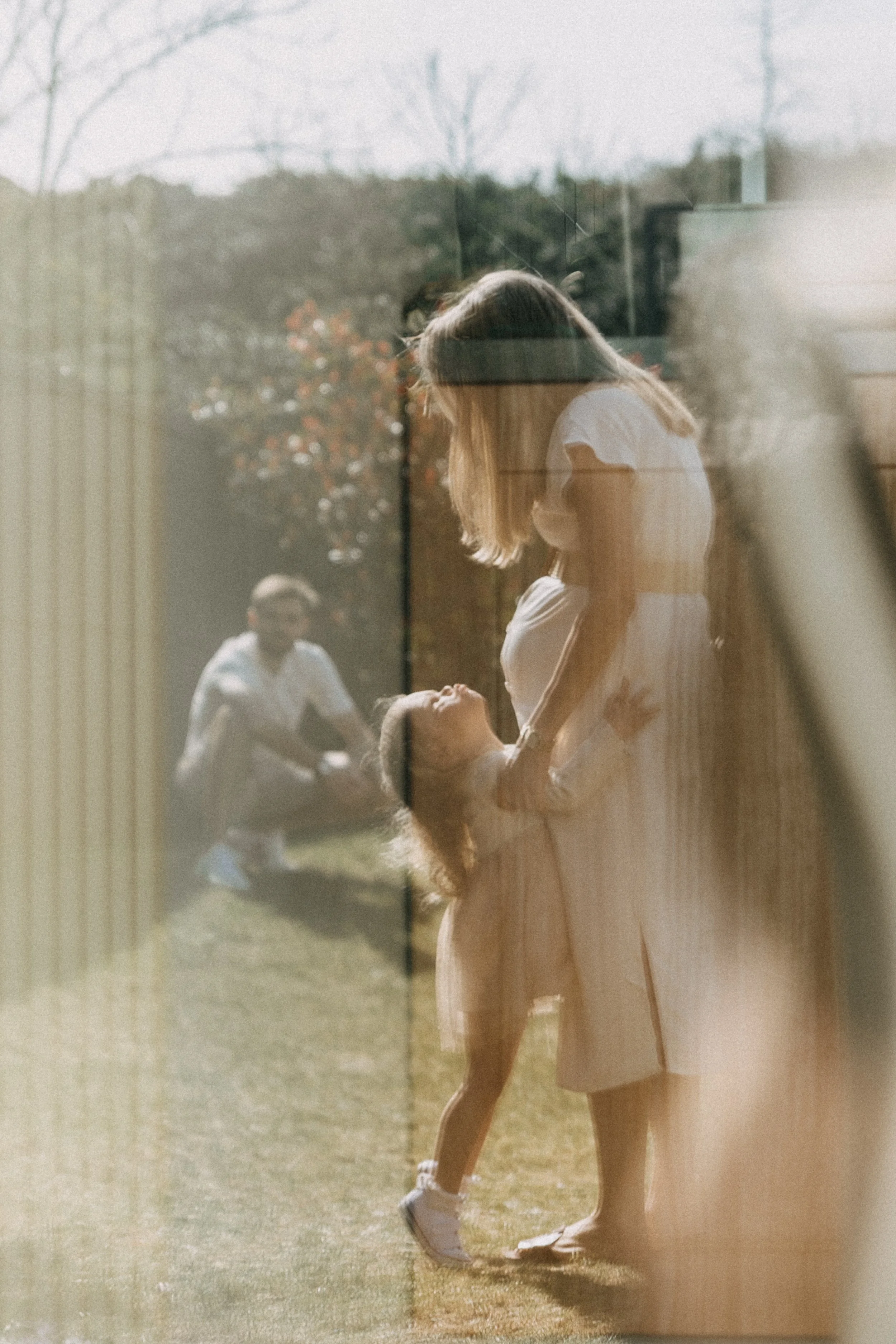 Une femme en robe blanche et une jeune fille regardent et se tiennent la main à l'extérieur, vus à travers une fenêtre, avec d'autres personnes en arrière-plan et un paysage naturel.