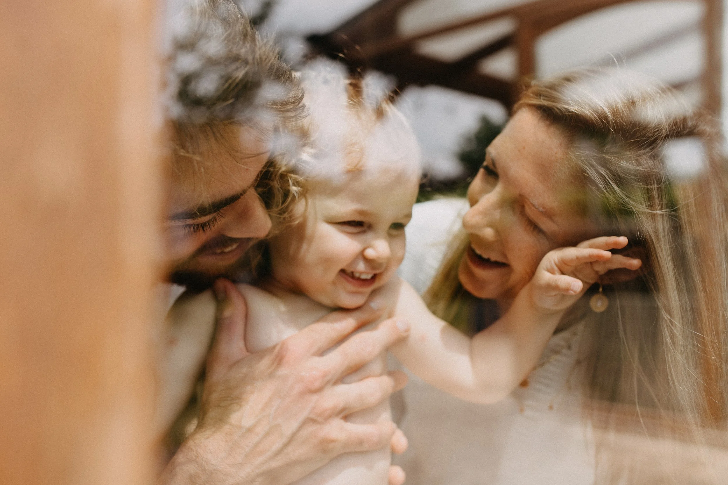 Une famille de trois personnes, un homme, une femme et un enfant, sourient et partagent un moment de joie ensemble à l'intérieur.