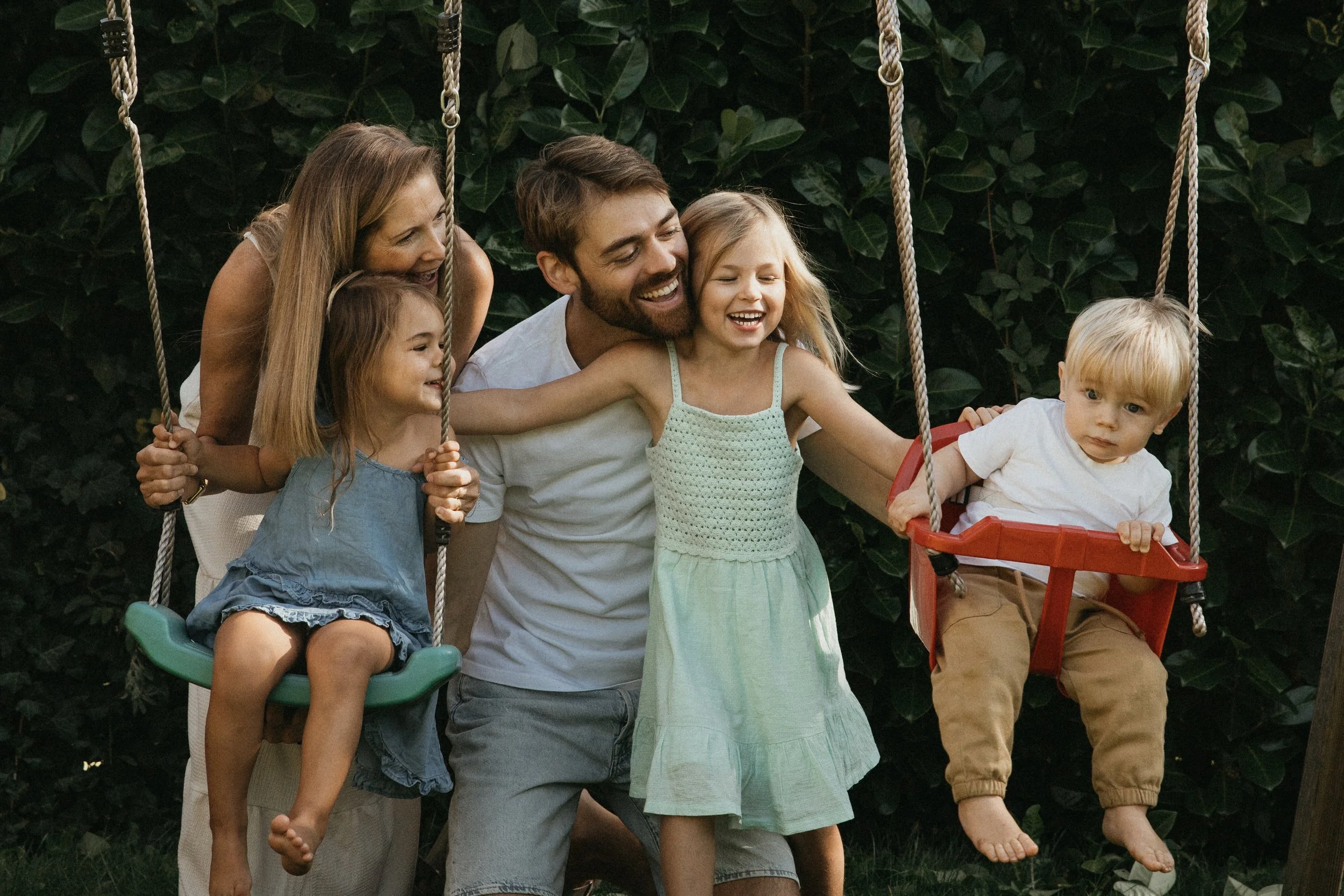 Une famille de cinq personnes joue dans un jardin avec des balançoires. Deux enfants sont assis dans des balançoires, une fille historiquement sur la gauche avec un t-shirt bleue et un garçon à droite avec un t-shirt blanc. Une mère, un père et une a