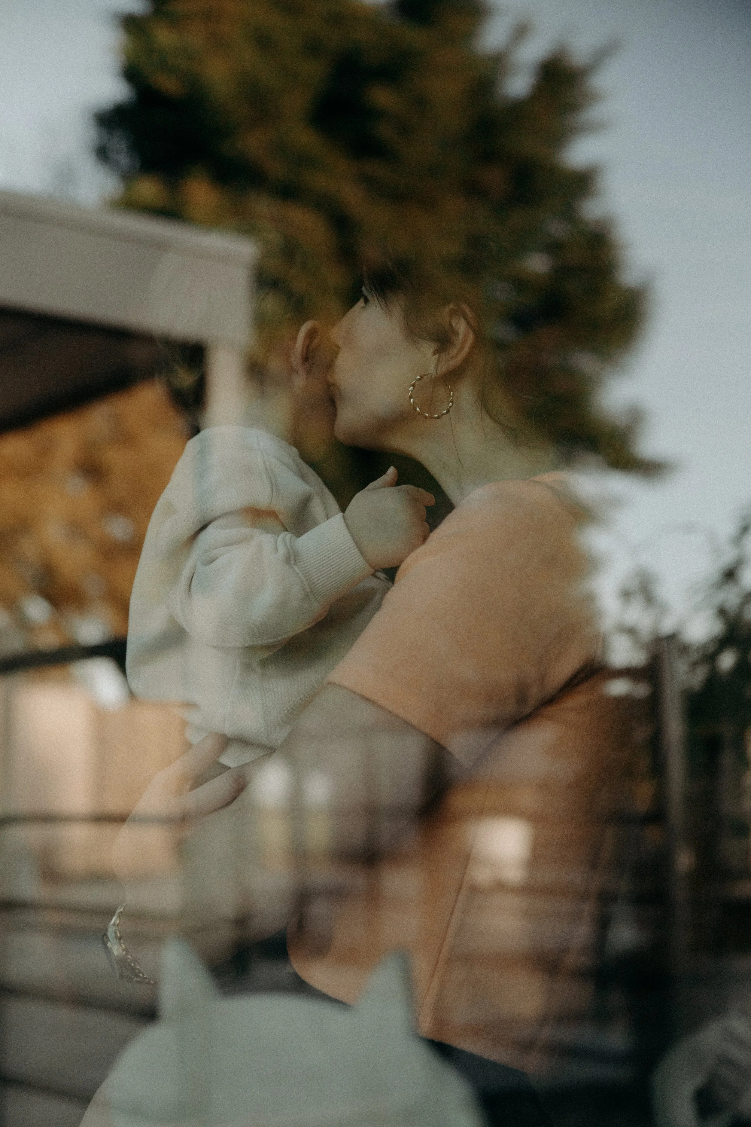 Une femme tenant un jeune enfant dans ses bras, vue à travers une vitre avec un reflet d'arbres et un chat en silhouette