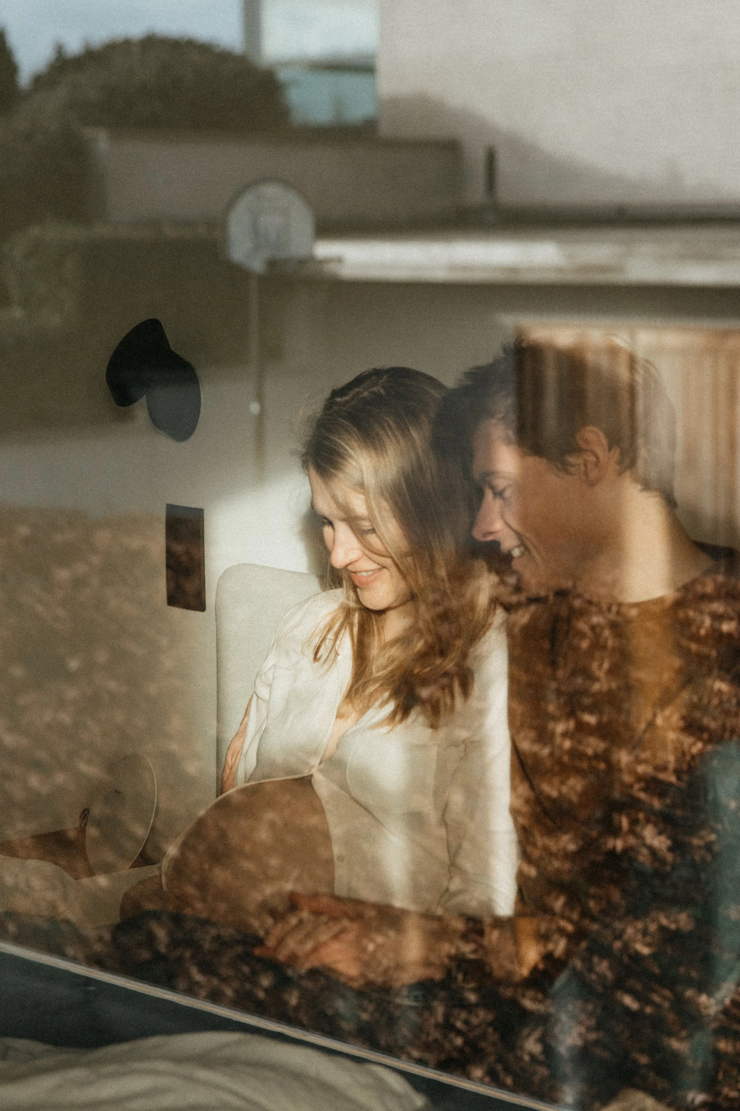 Une femme et un homme souriants regardant par une fenêtre, avec une réflexion de leur intérieur et d'un paysage extérieur dans le verre.