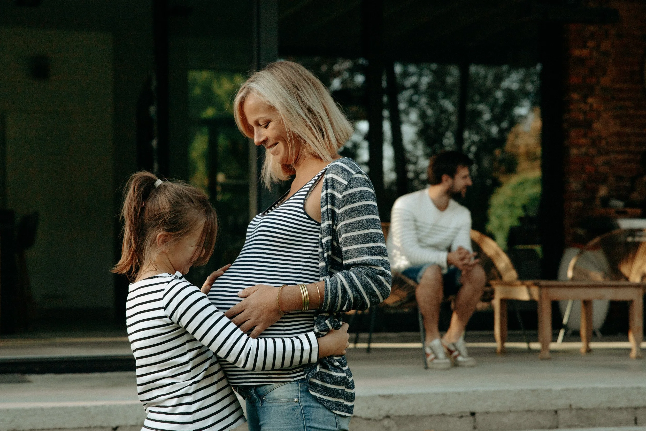 Une femme enceinte et une jeune fille s'embrassent affectueusement, tandis qu'un homme, assis dehors en arrière-plan, les regarde sourire, dans un cadre extérieur chaleureux.