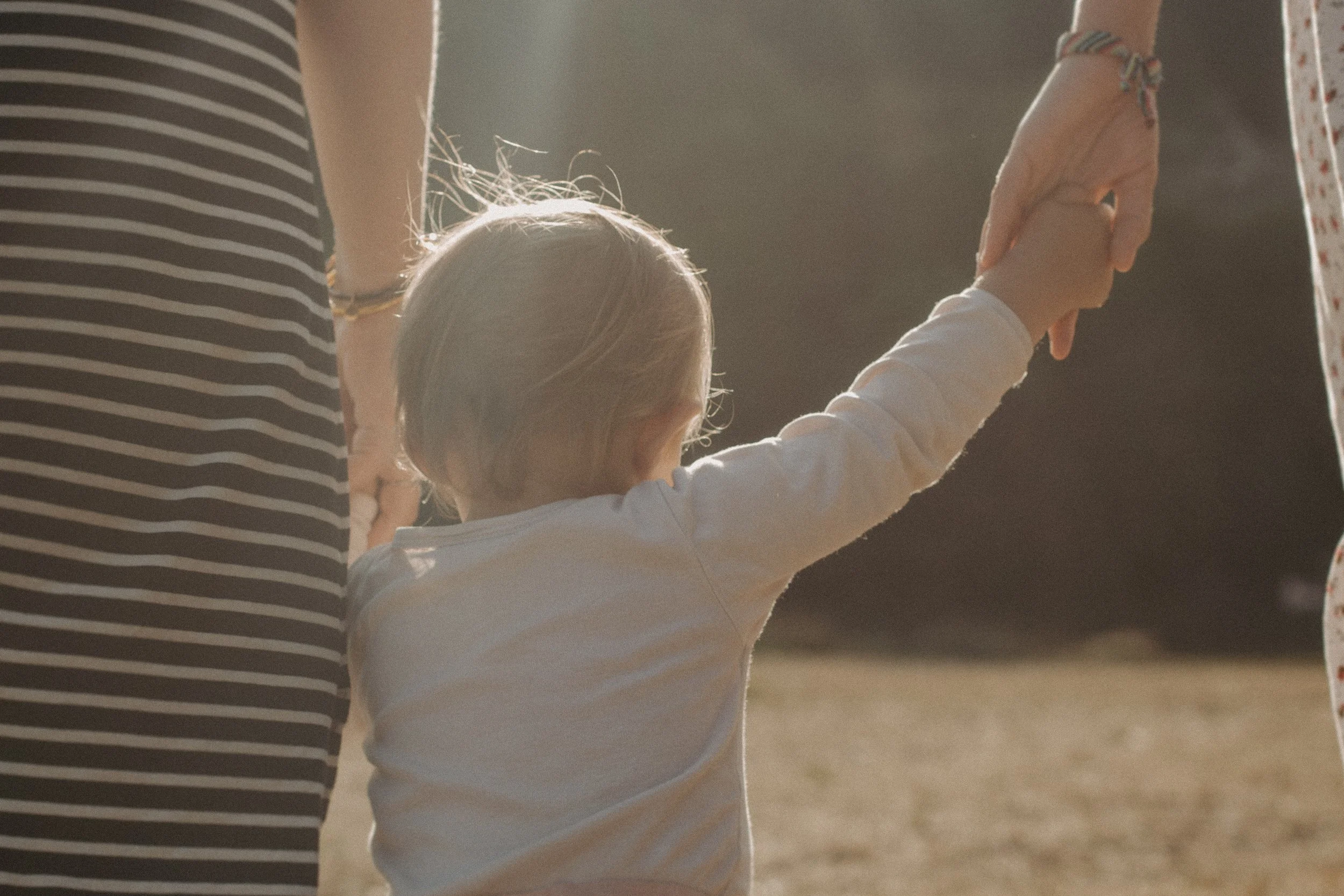 Un enfant en blanc tenant la main d'une adulte en chemise blanche à pois rouges dans un environnement en plein air avec un soleil chaud.