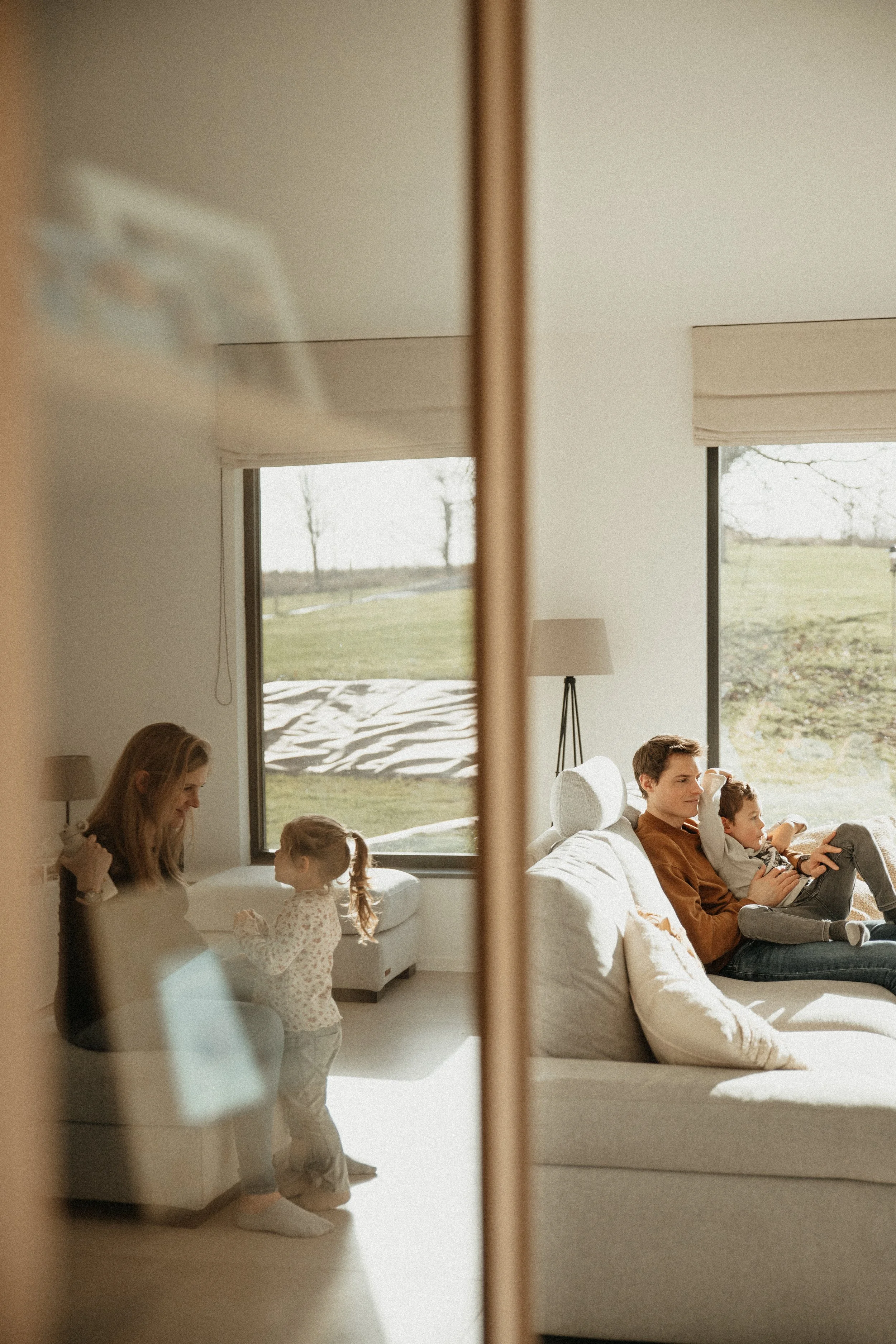 Une famille de quatre personnes dans un salon, vue à travers une porte ou un miroir, avec deux grandes fenêtres offrant une vue sur l'extérieur ensoleillé.