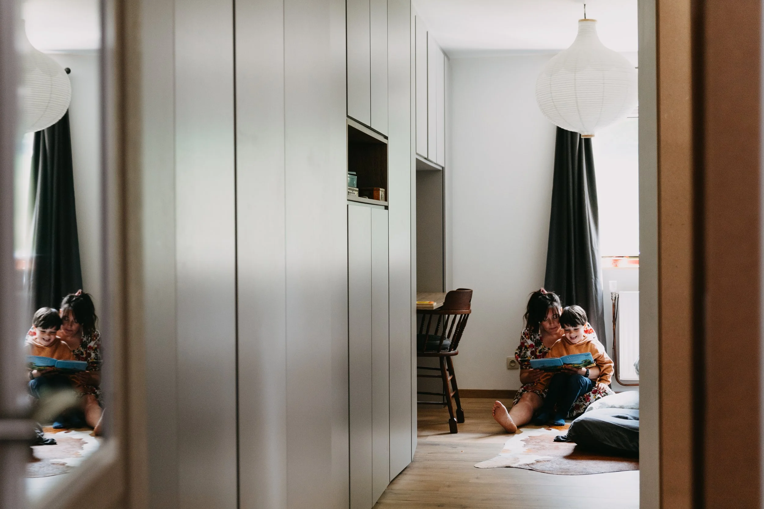 Une mère et son enfant assis sur un tapis à lire un livre dans une pièce ensoleillée. La pièce a des murs blancs, un placard gris, une chaise en bois, et une fenêtre avec des rideaux noirs.