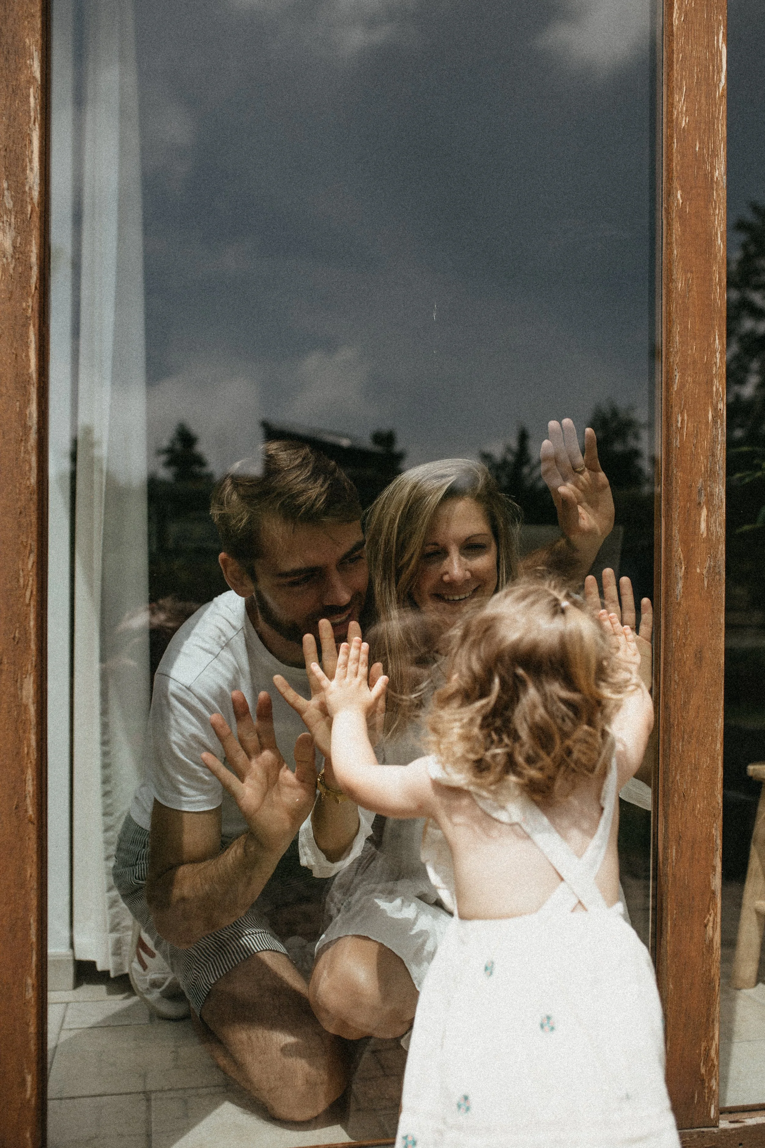 Un couple et une enfant jouent contre une vitre, leur reflet étant visible, dans un environnement intérieur avec un extérieur visible.