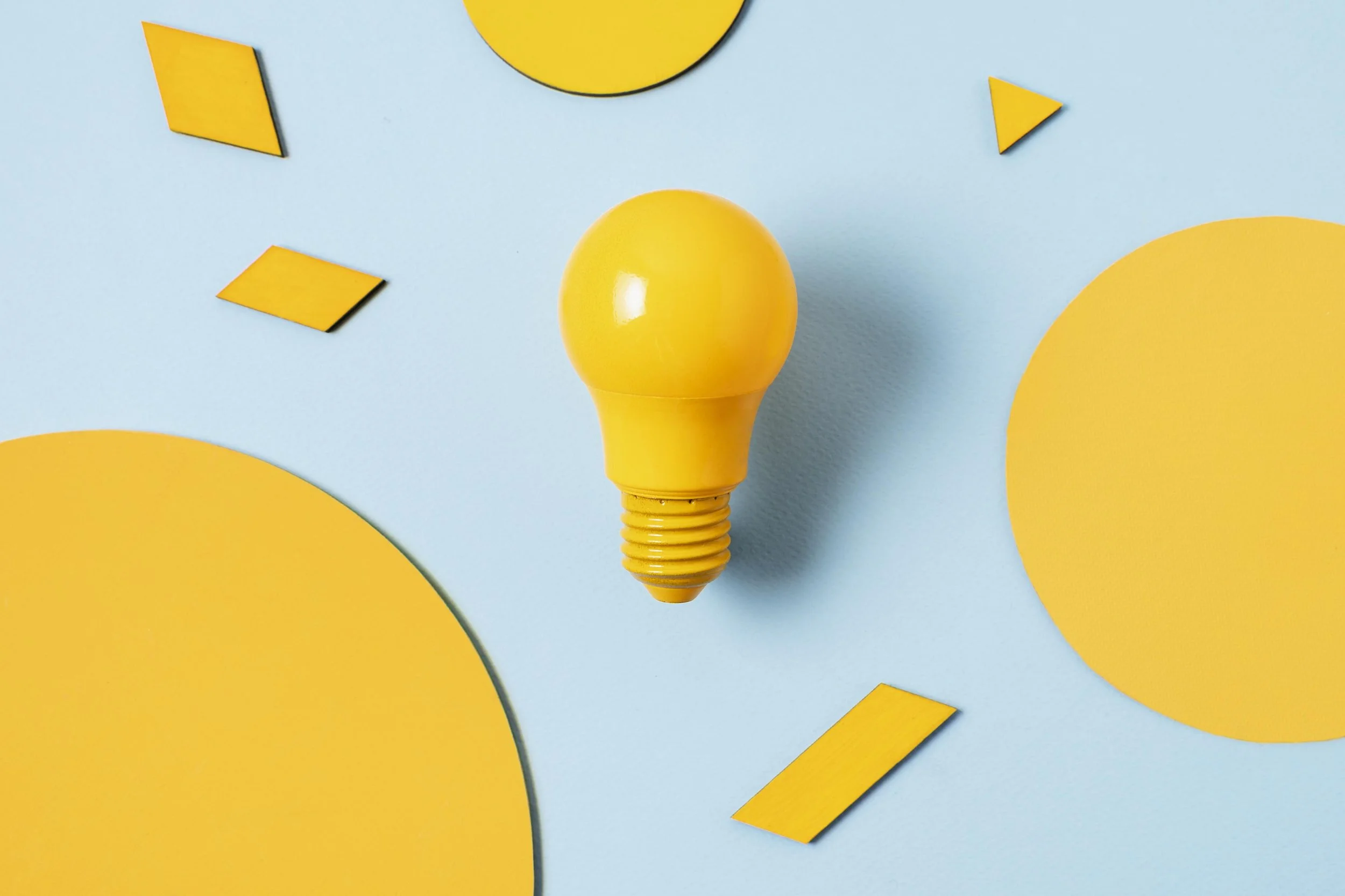 A yellow light bulb-shaped object on a light blue surface, surrounded by abstract yellow shapes and geometric pieces.