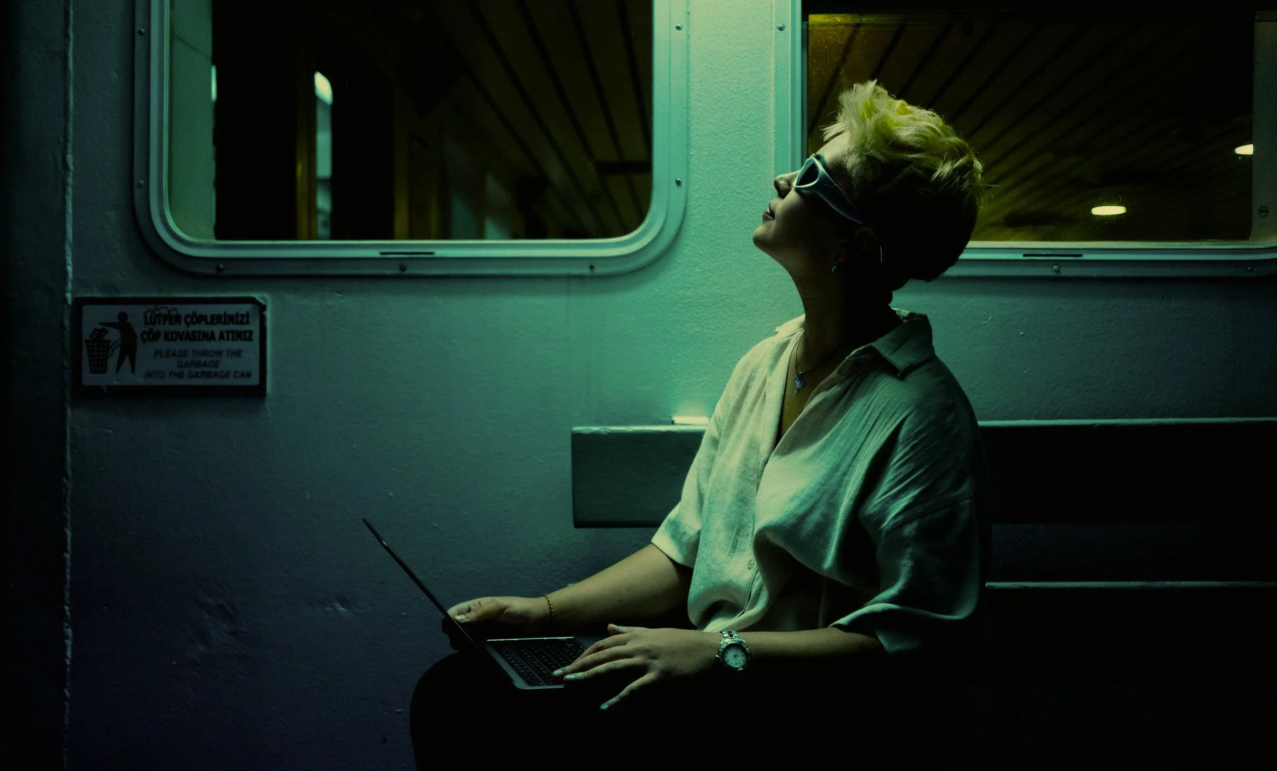 A woman with short blond hair, wearing sunglasses and a beige shirt, sits with her eyes closed and head tilted back next to a window at night, holding a laptop on her lap.