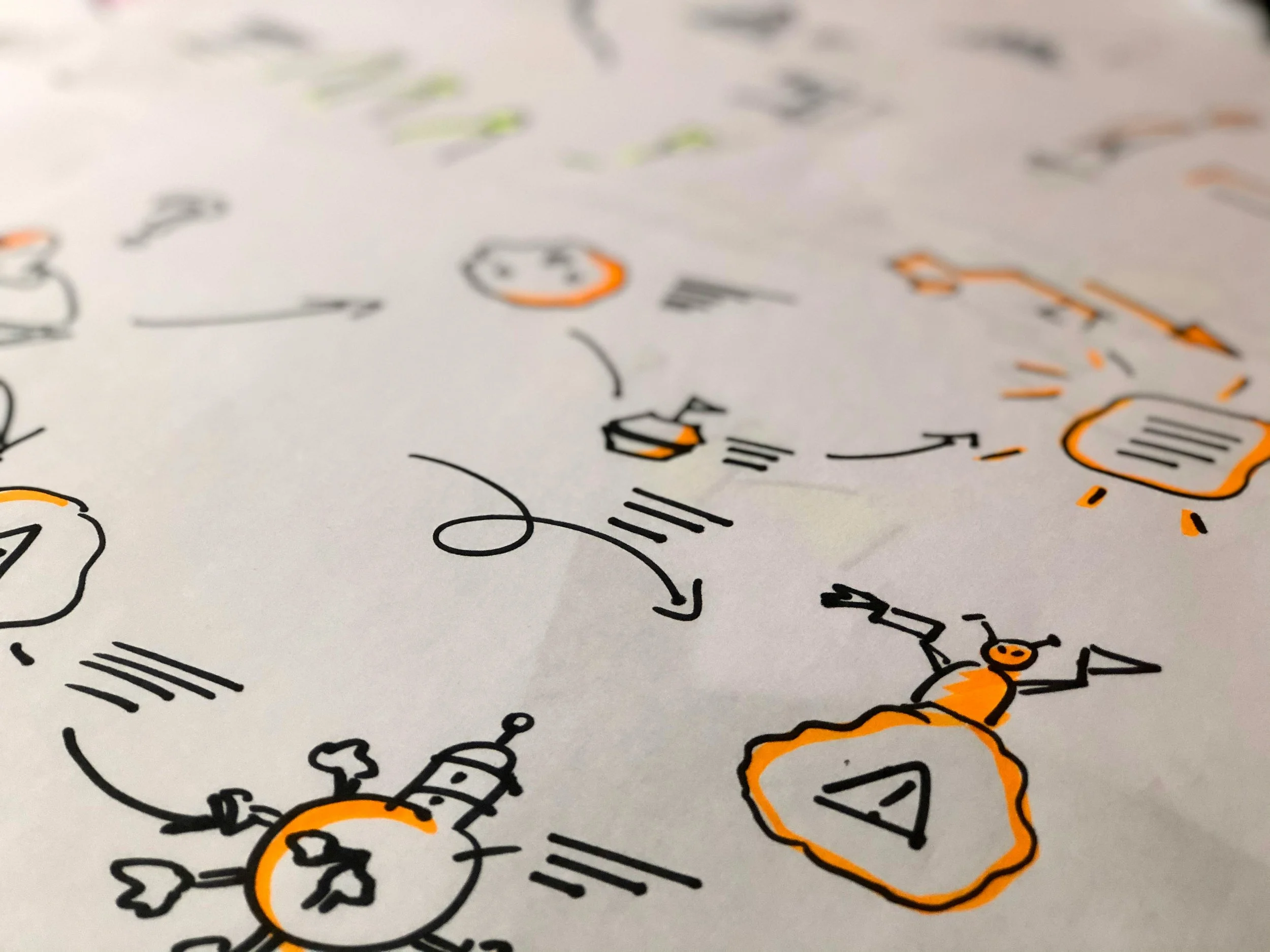 Close-up of a sheet of paper with drawings and handwritten notes in black marker and orange highlights, depicting diagrams, icons, and arrows.