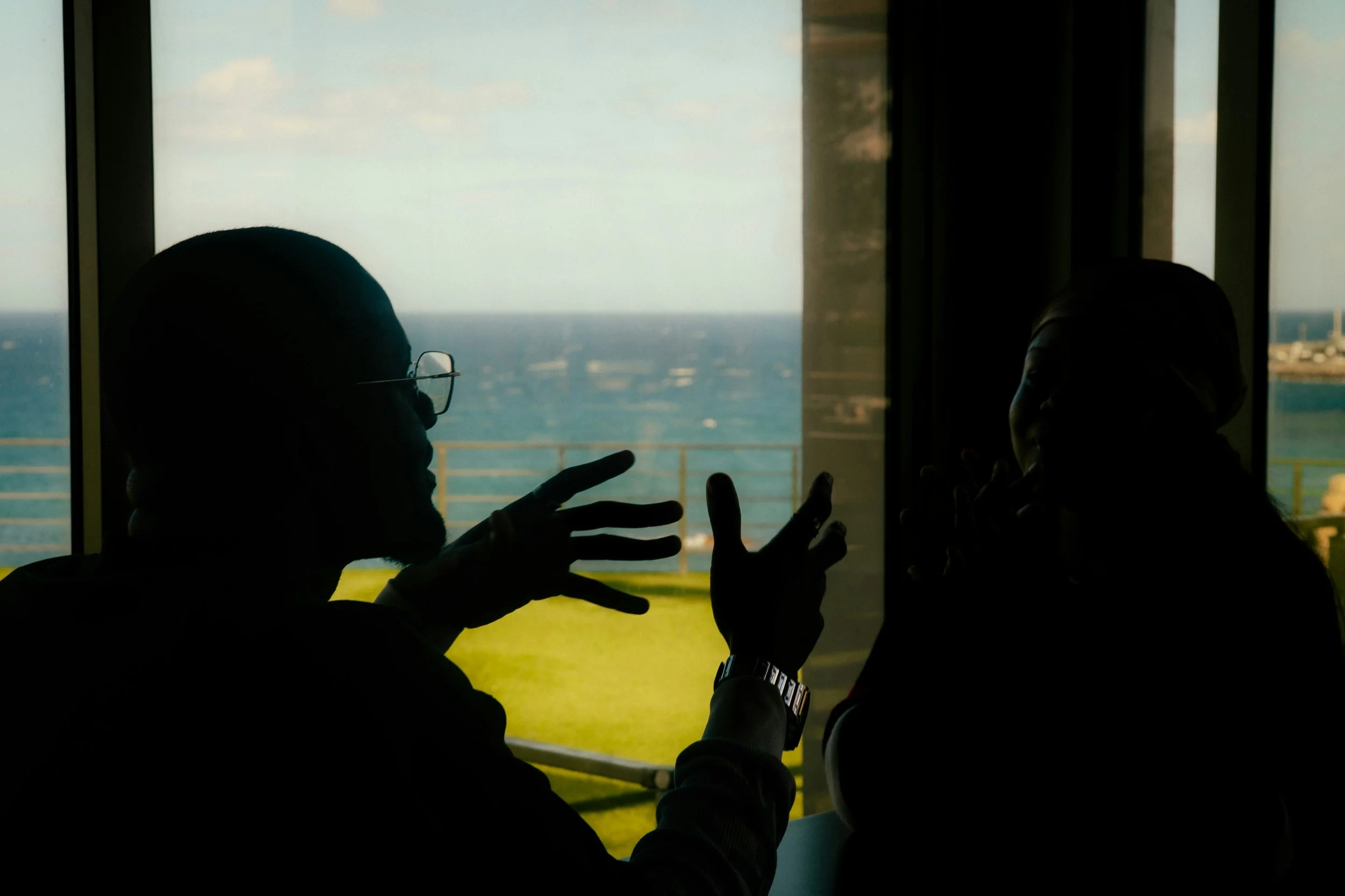 Silhouettes of two people talking in front of a large window overlooking a body of water and a distant shoreline.
