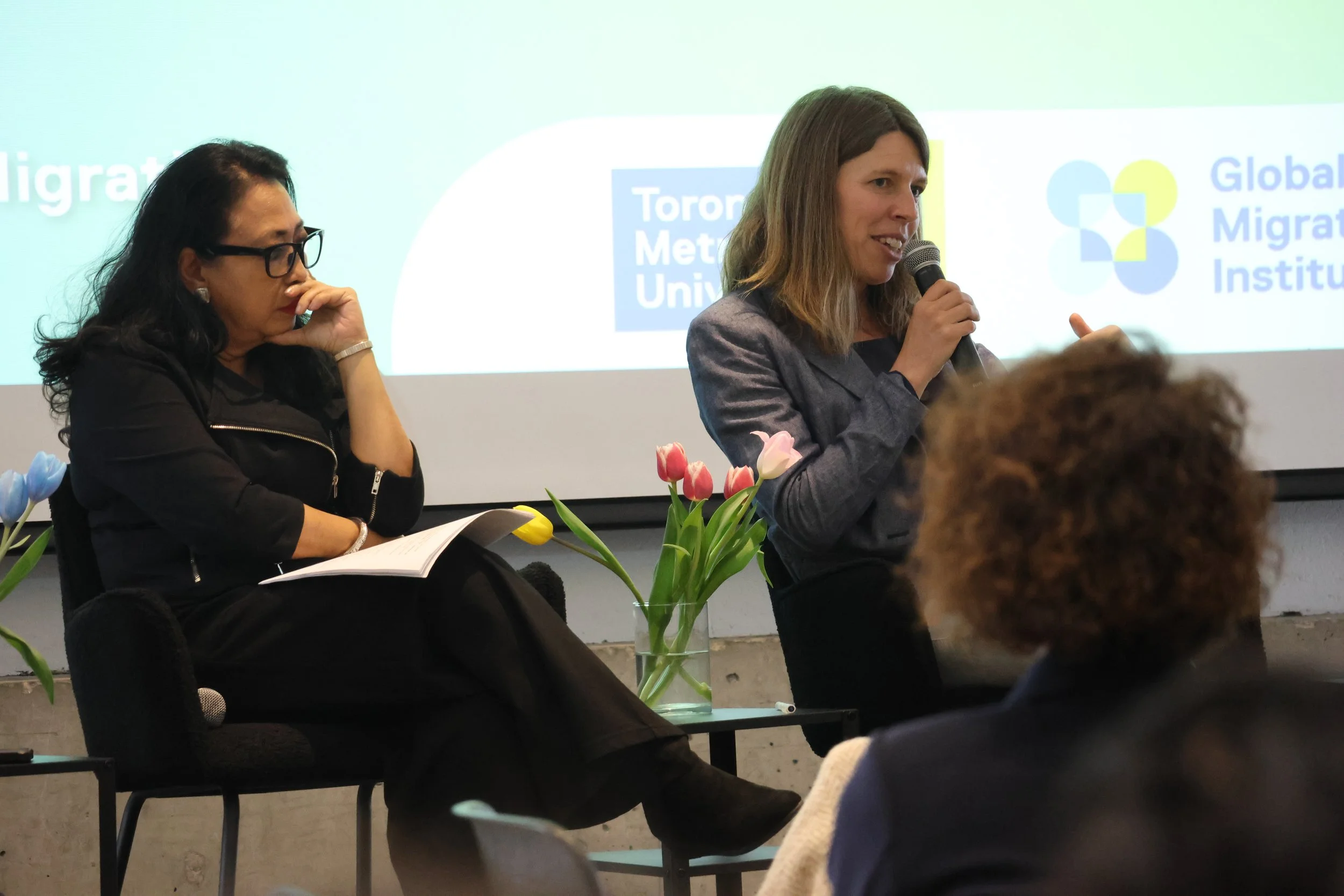 Two women participating in a panel discussion at an event, with a screen displaying logos of Toronto Metropolitan University and the Global Migration Institute in the background. There are pink and white tulips on the table.